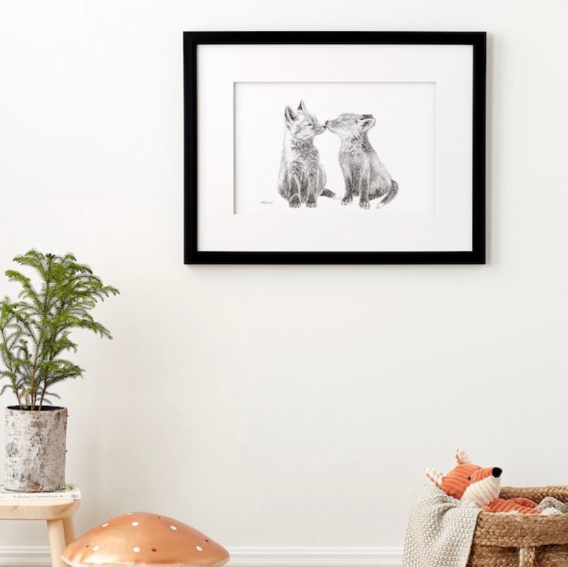 Framed artwork of two foxes on a wall with a plant and toy in the foreground.
