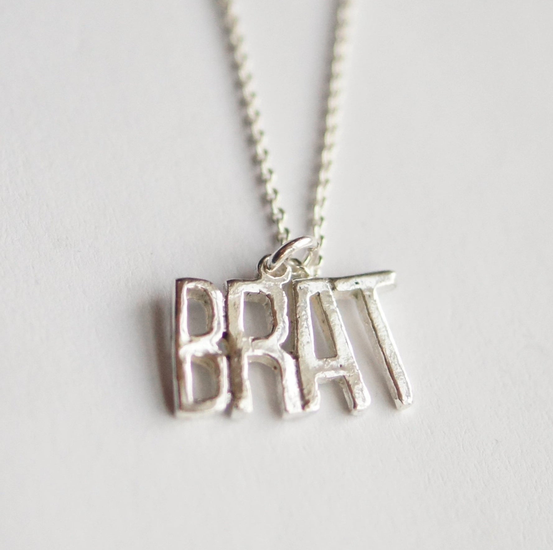 Silver necklace with 'BRAT' pendant on a light gray background