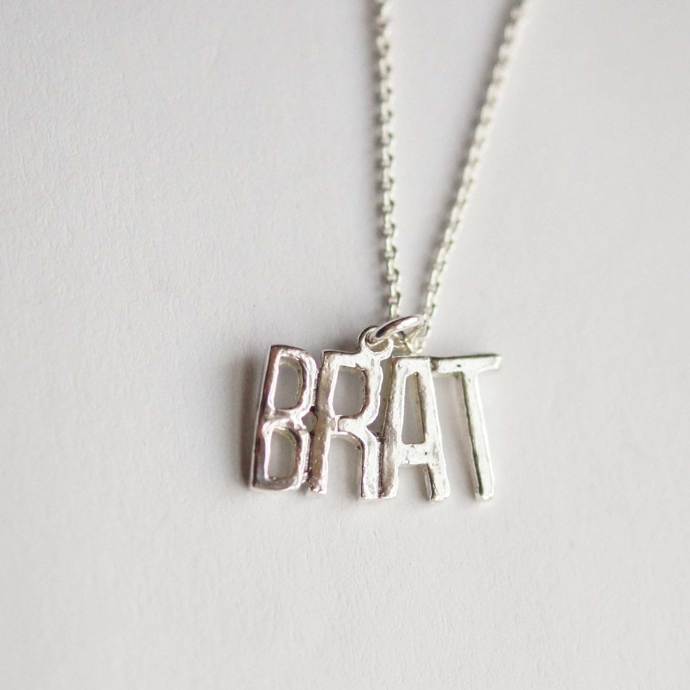 Silver necklace with 'BRAT' pendant on a light gray background