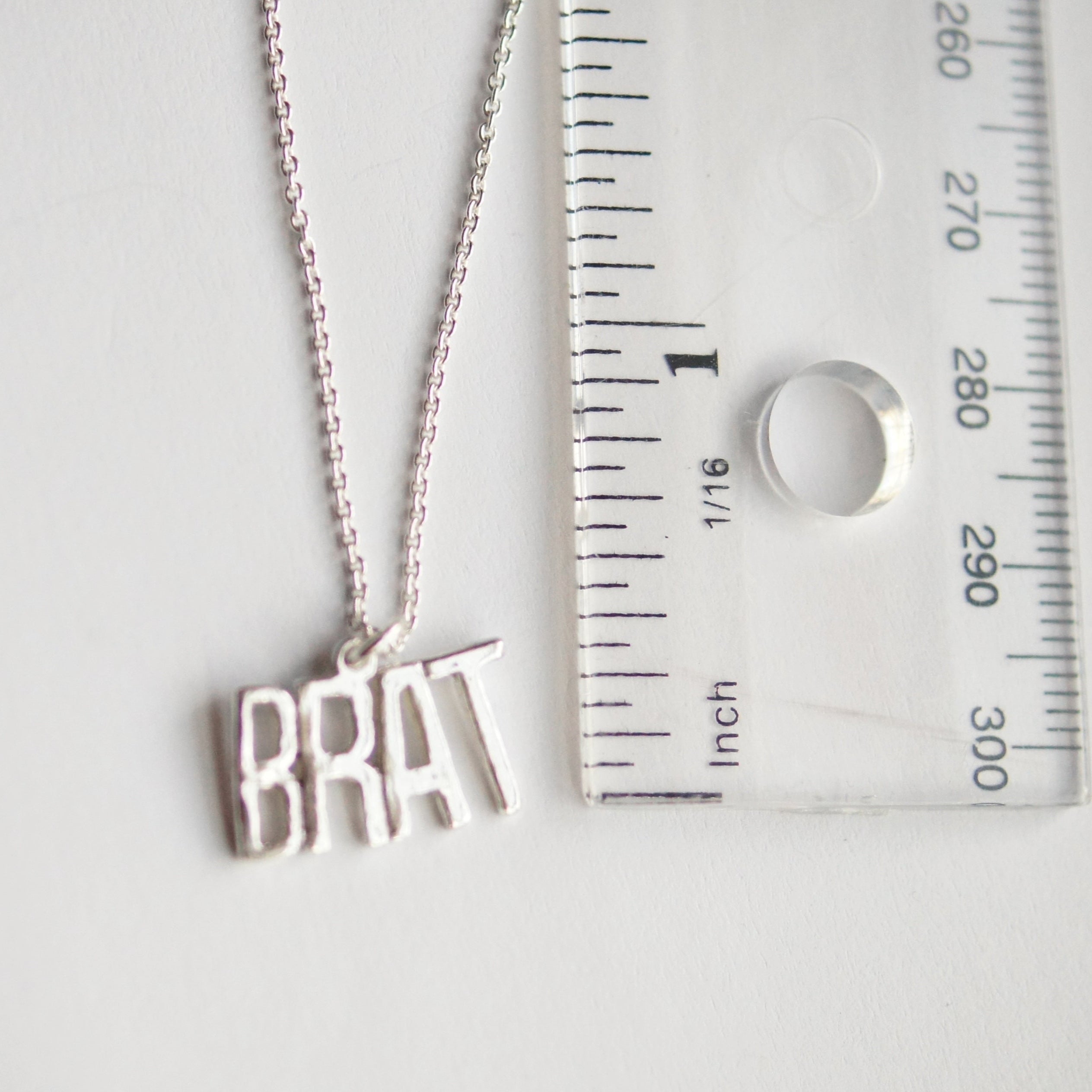 Silver necklace with 'BRAT' pendant next to a ruler for size reference on a white background