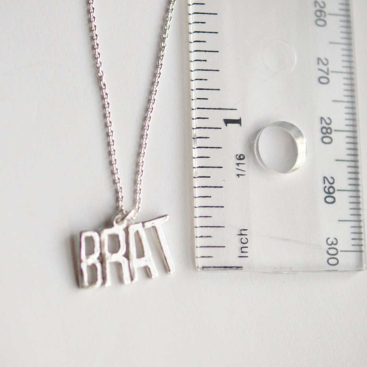 Silver necklace with 'BRAT' pendant next to a ruler for size reference on a white background