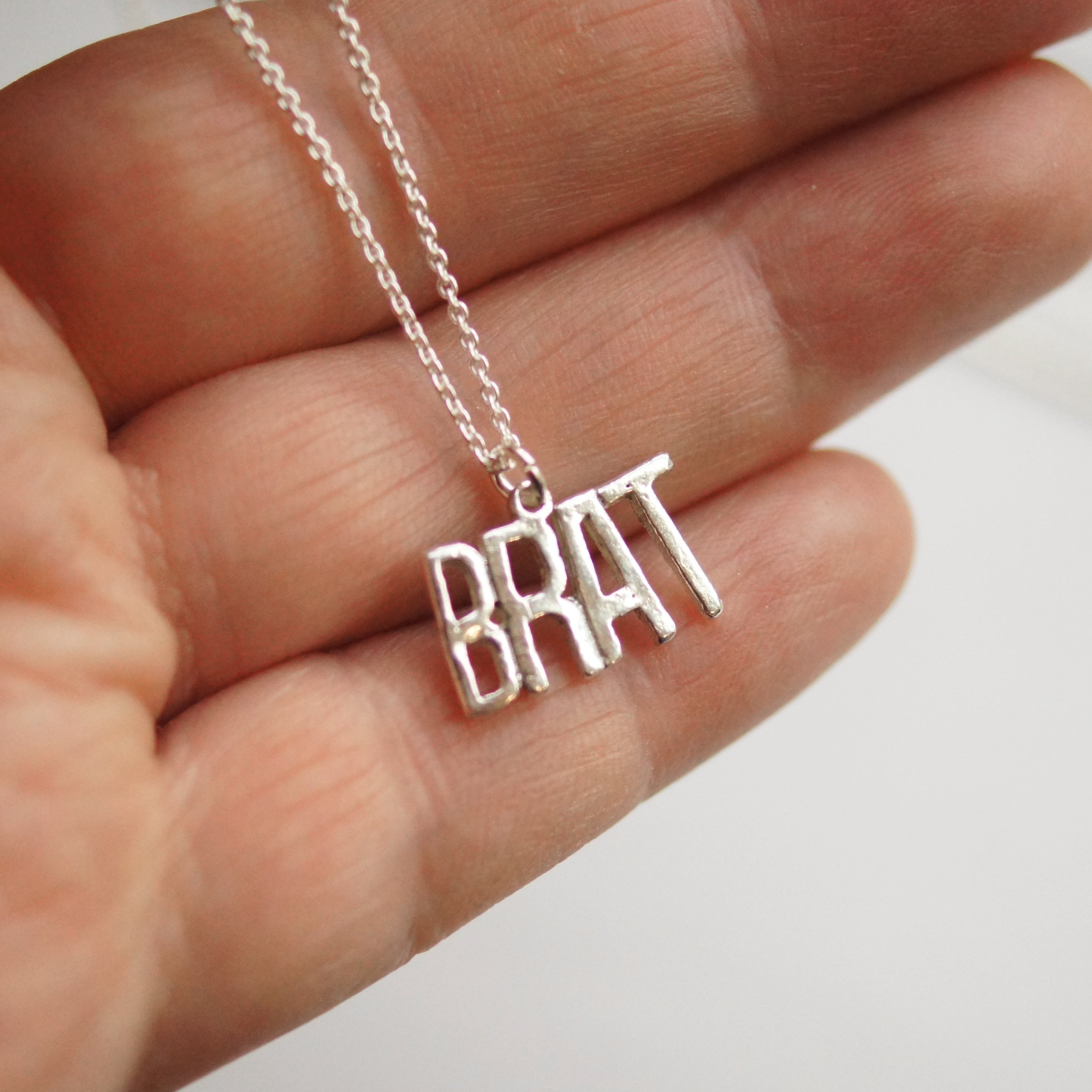 Silver necklace with 'BRAT' pendant held in a hand against a neutral background