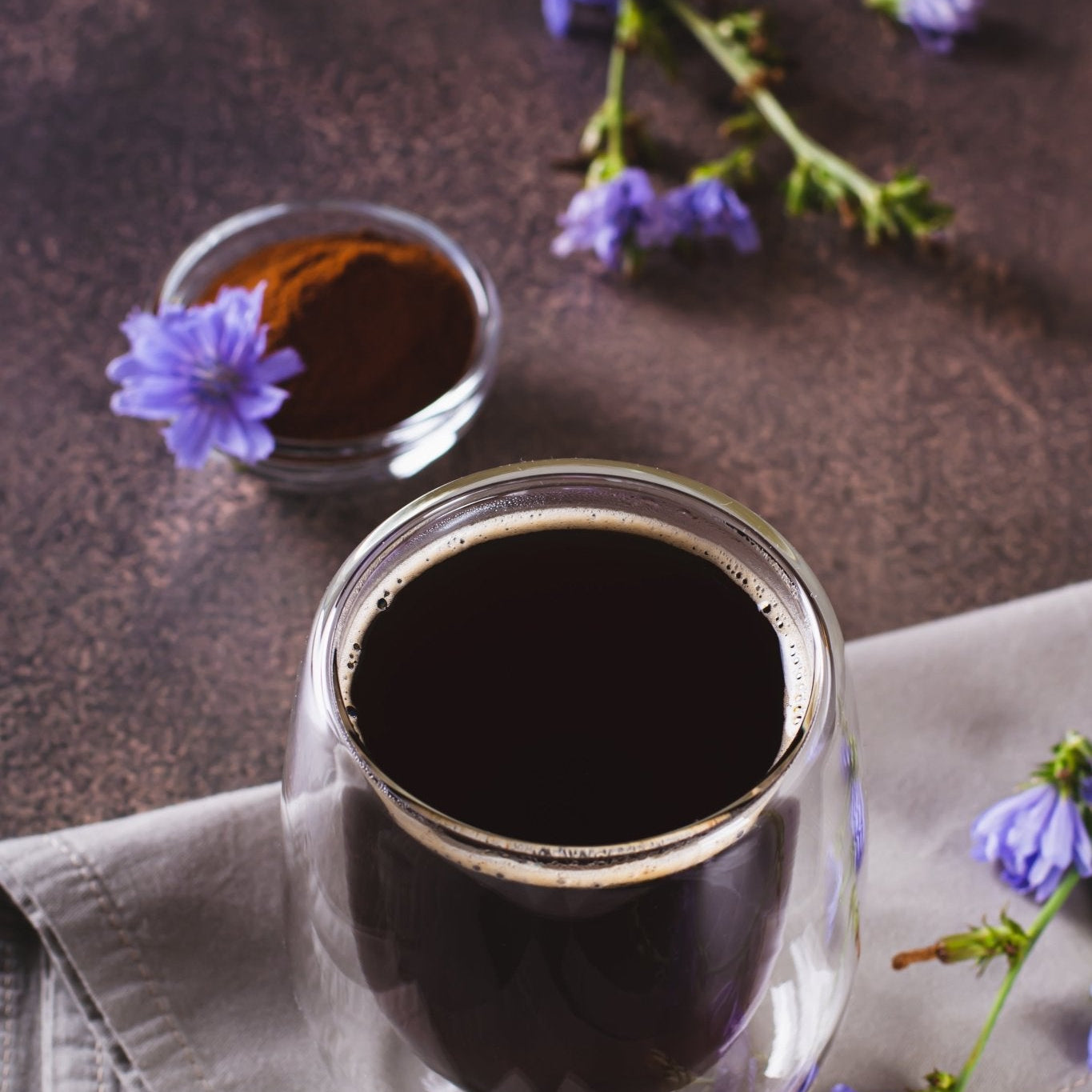 Glass jar of dark liquid with a small container of liquid and purple flowers on a brown surface.