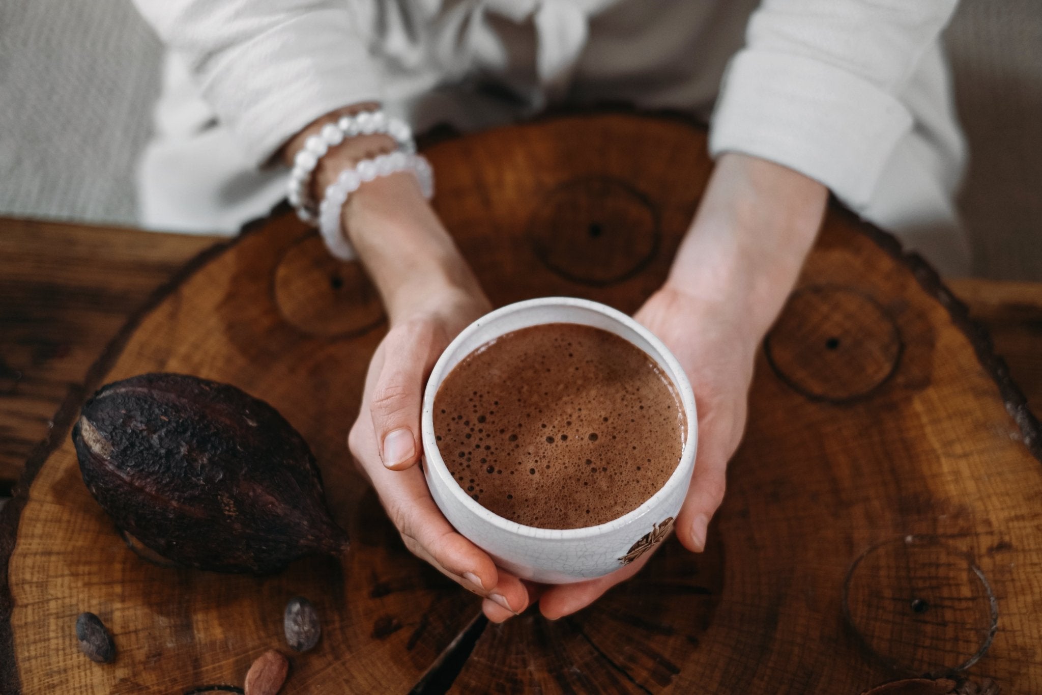 Person holding a cup of hot chocolate on a wooden surface with cocoa beans.