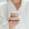 Person holding a small, round, beige container with a textured surface against a blurred background