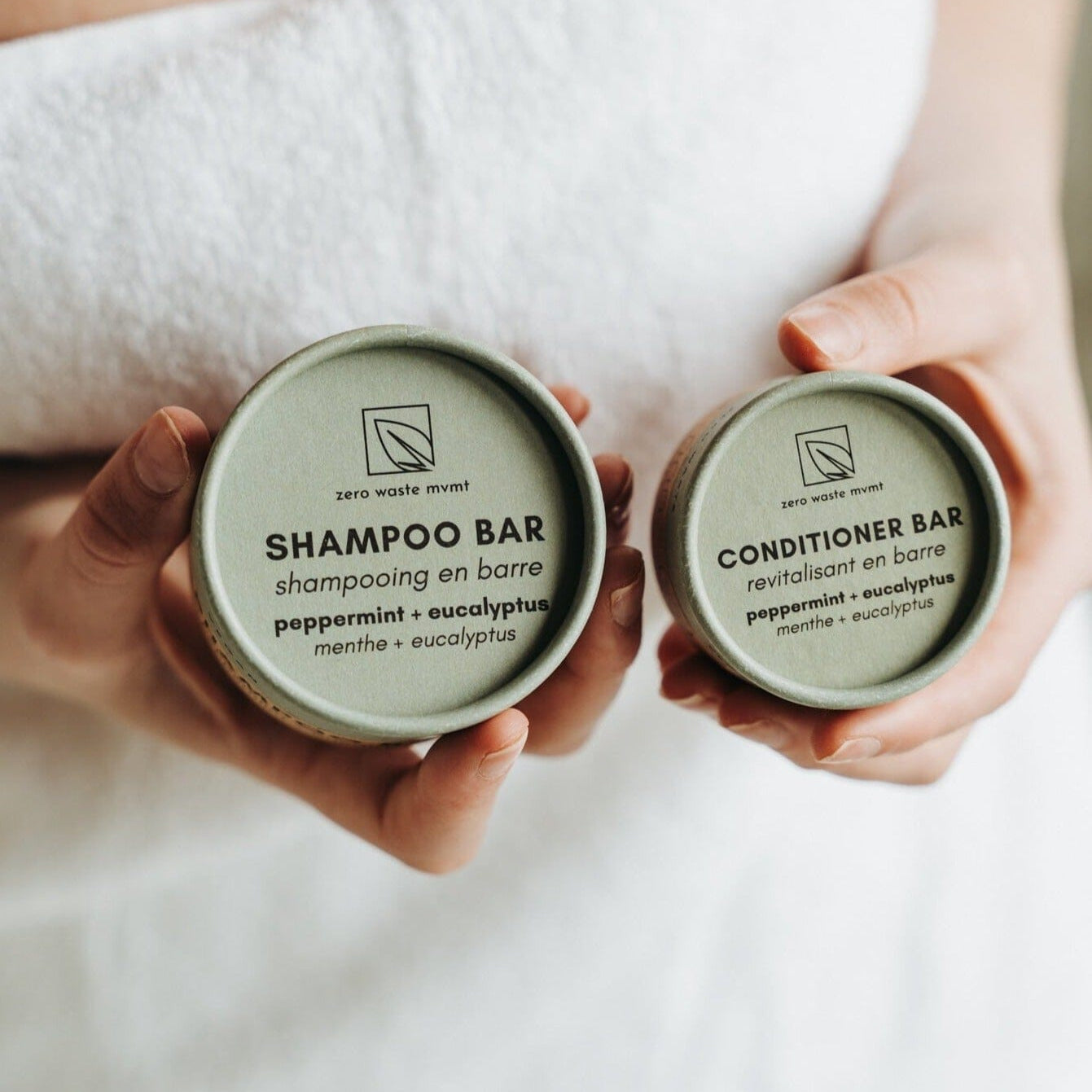 Two hands holding containers labeled 'Shampoo Bar' and 'Conditioner Bar' with a neutral background.