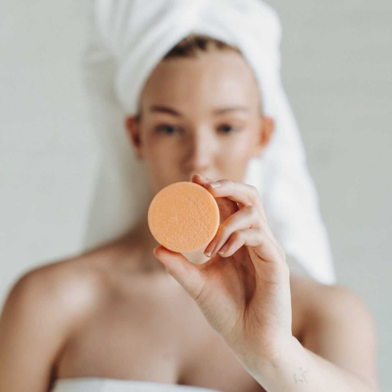 Person holding a beauty sponge with a towel on their head against a neutral background