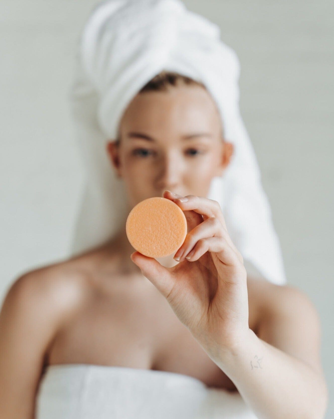 Person holding a beauty sponge with a towel on their head against a neutral background