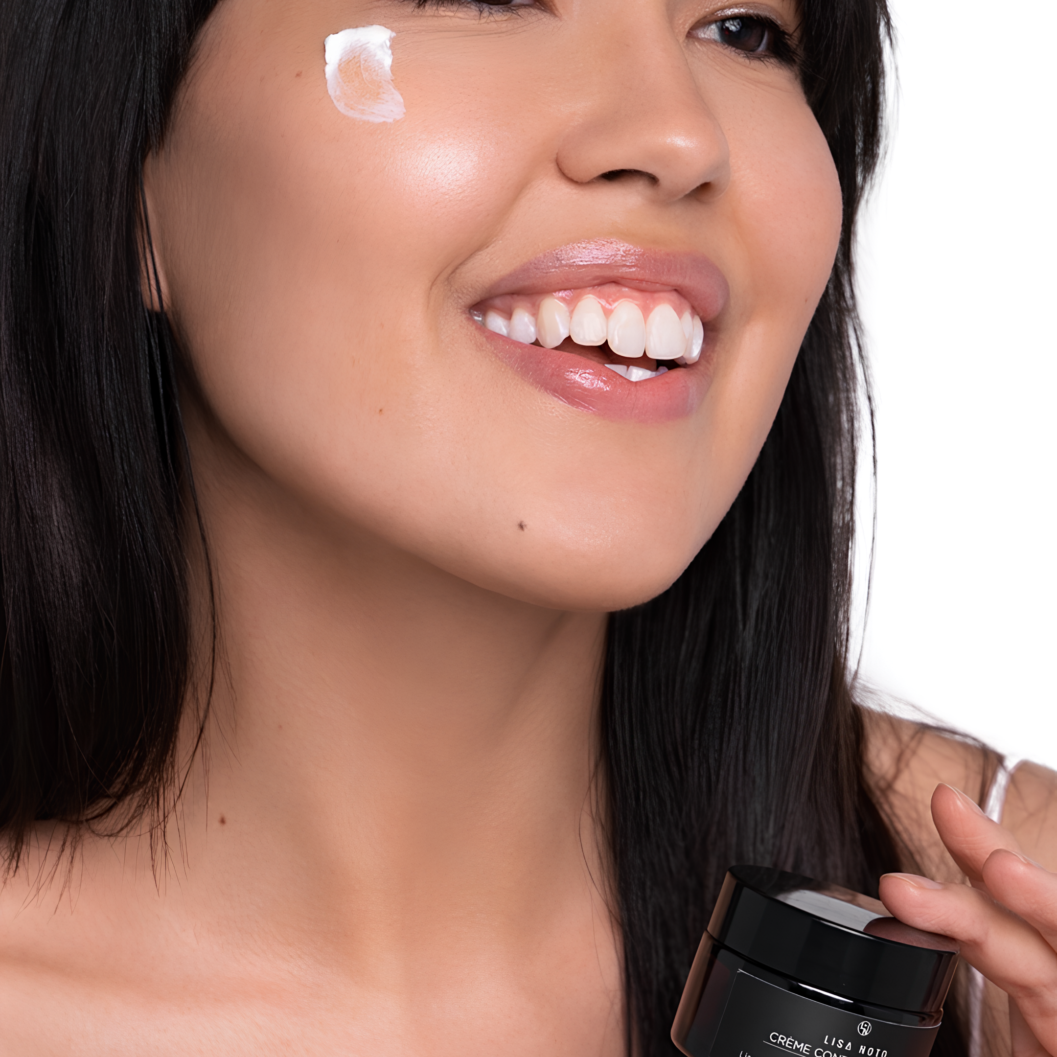 Close-up of a smiling woman with white cream on her cheek, holding a black jar of Lisa Noto eye contour cream 