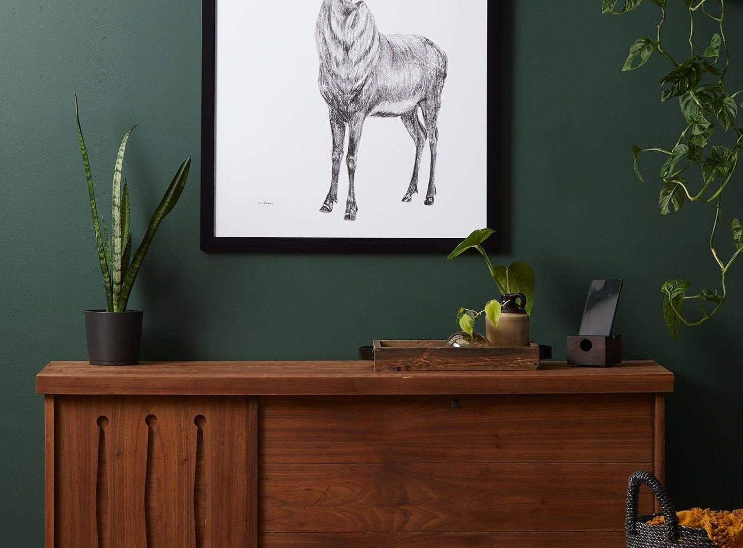 Wooden sideboard against a green wall with a framed deer print above it, surrounded by plants.