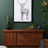 Wooden sideboard against a green wall with a framed deer print above it, surrounded by plants.