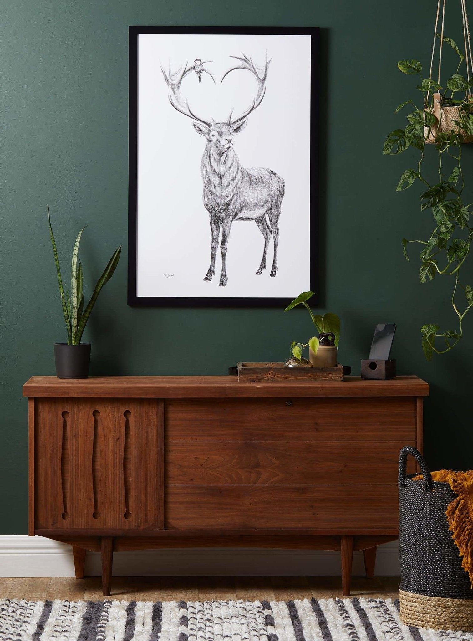 Wooden sideboard against a green wall with a framed deer print above it, surrounded by plants.