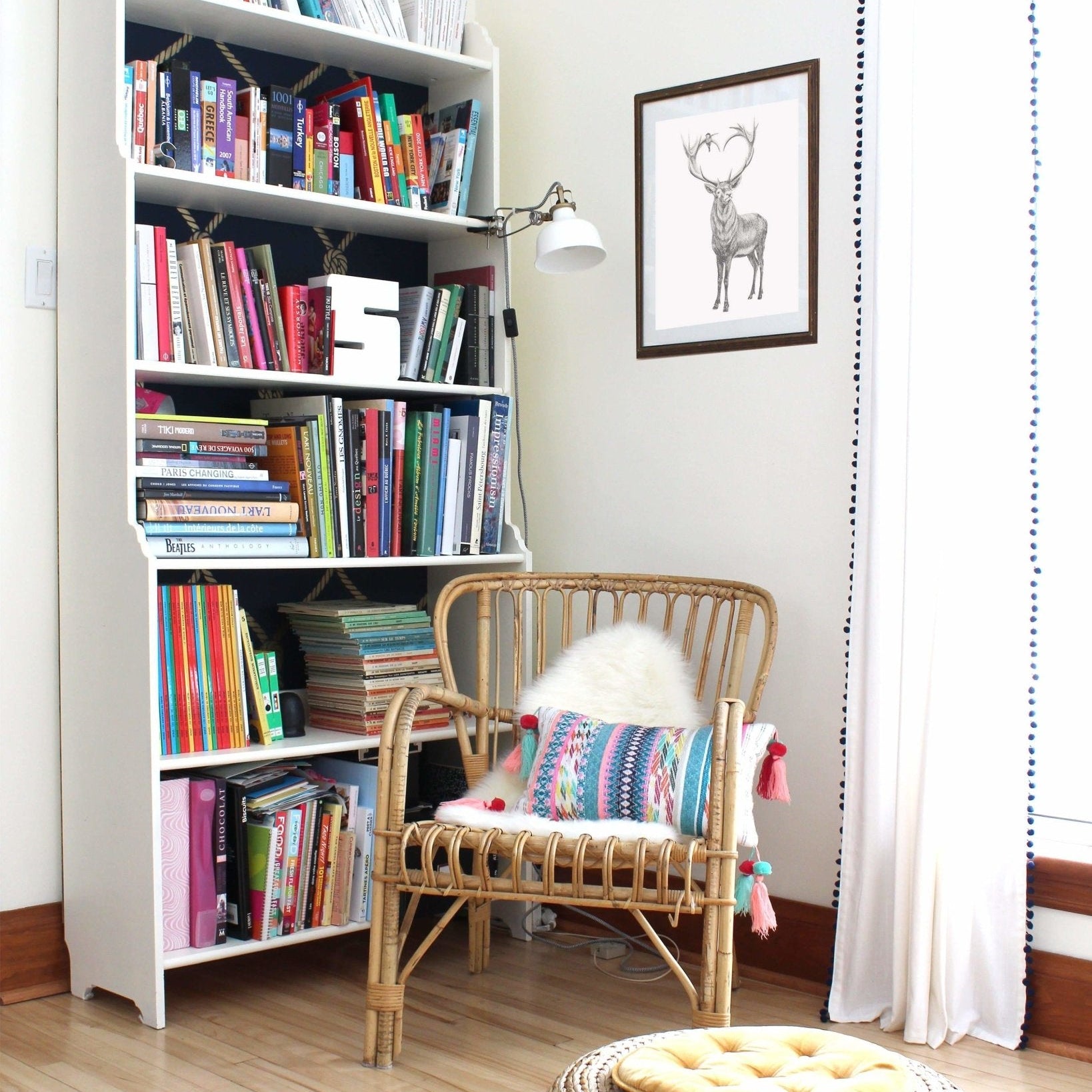 White bookshelf filled with books next to a wicker chair with a colorful pillow in a room.
