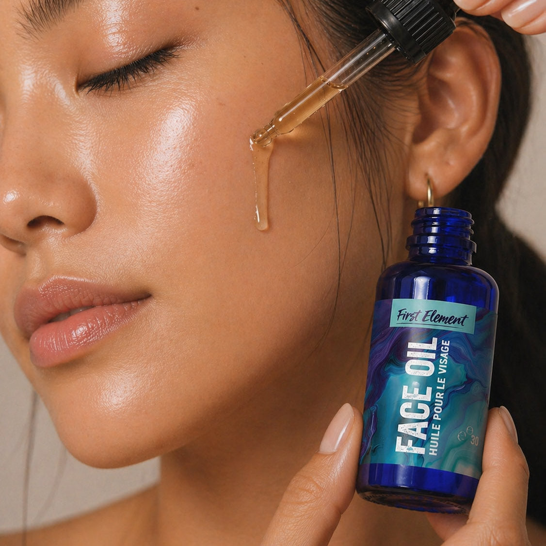 A close-up of a woman applying face oil with a dropper, holding a blue bottle labeled 'First Element Face Oil' near her cheek.