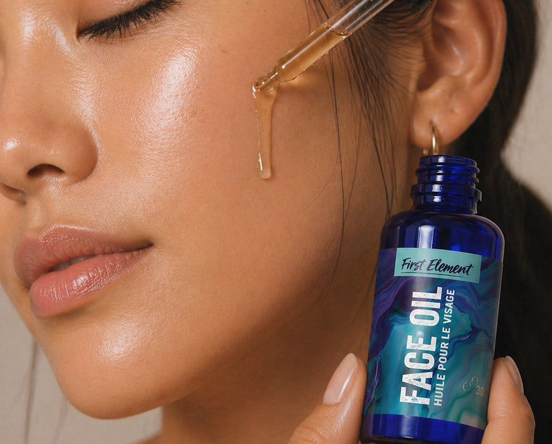 A close-up of a woman applying face oil with a dropper, holding a blue bottle labeled 'First Element Face Oil' near her cheek.