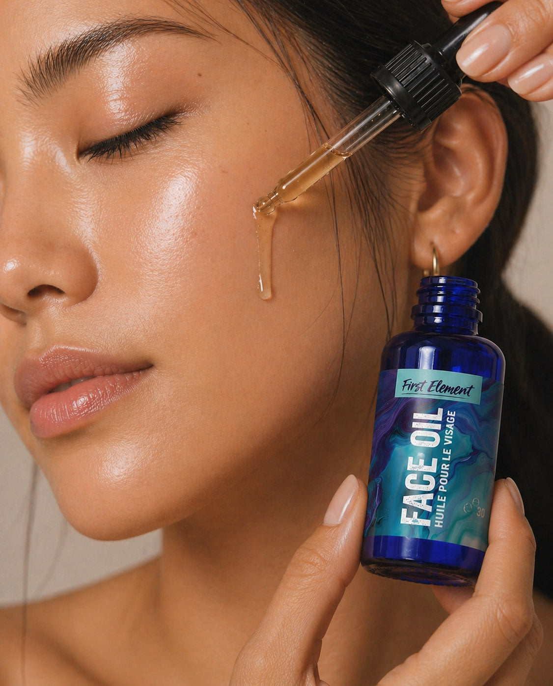 A close-up of a woman applying face oil with a dropper, holding a blue bottle labeled 'First Element Face Oil' near her cheek.