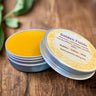 Yellow balm in a tin with a visible brand label on a wooden surface Kootenay Woman