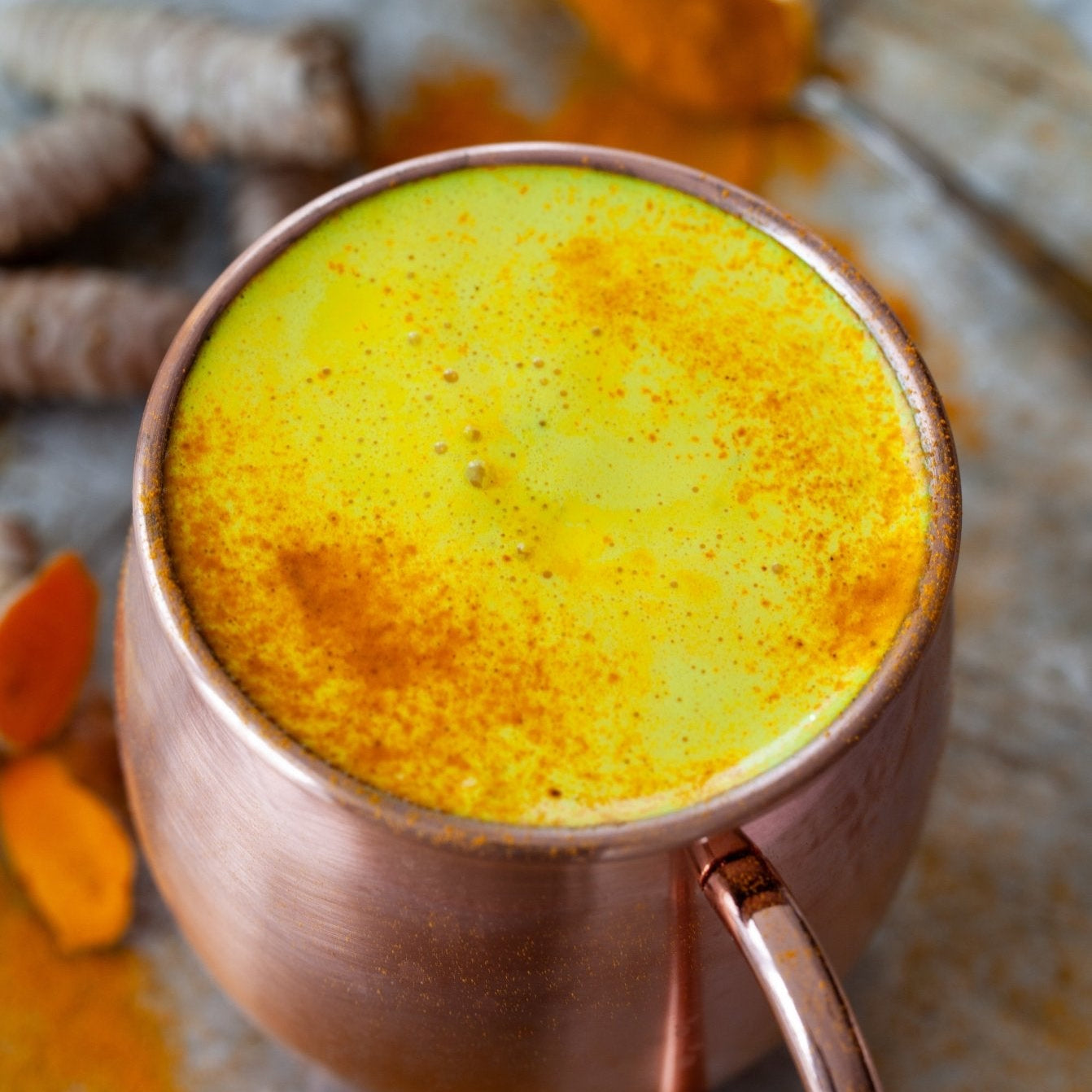 Copper mug filled with a yellow turmeric latte, surrounded by turmeric roots on a stone surface.