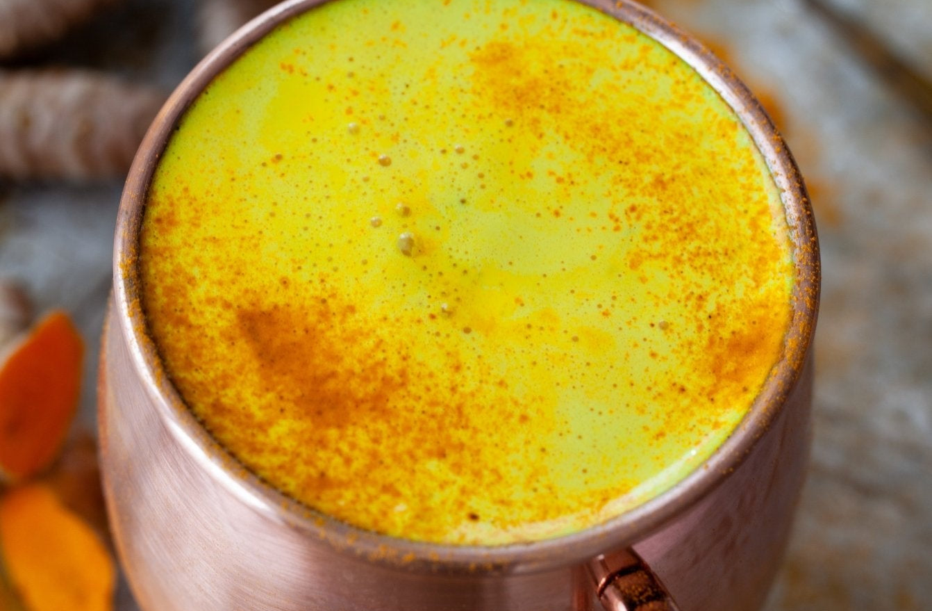 Copper mug filled with a yellow turmeric latte, surrounded by turmeric roots on a stone surface.