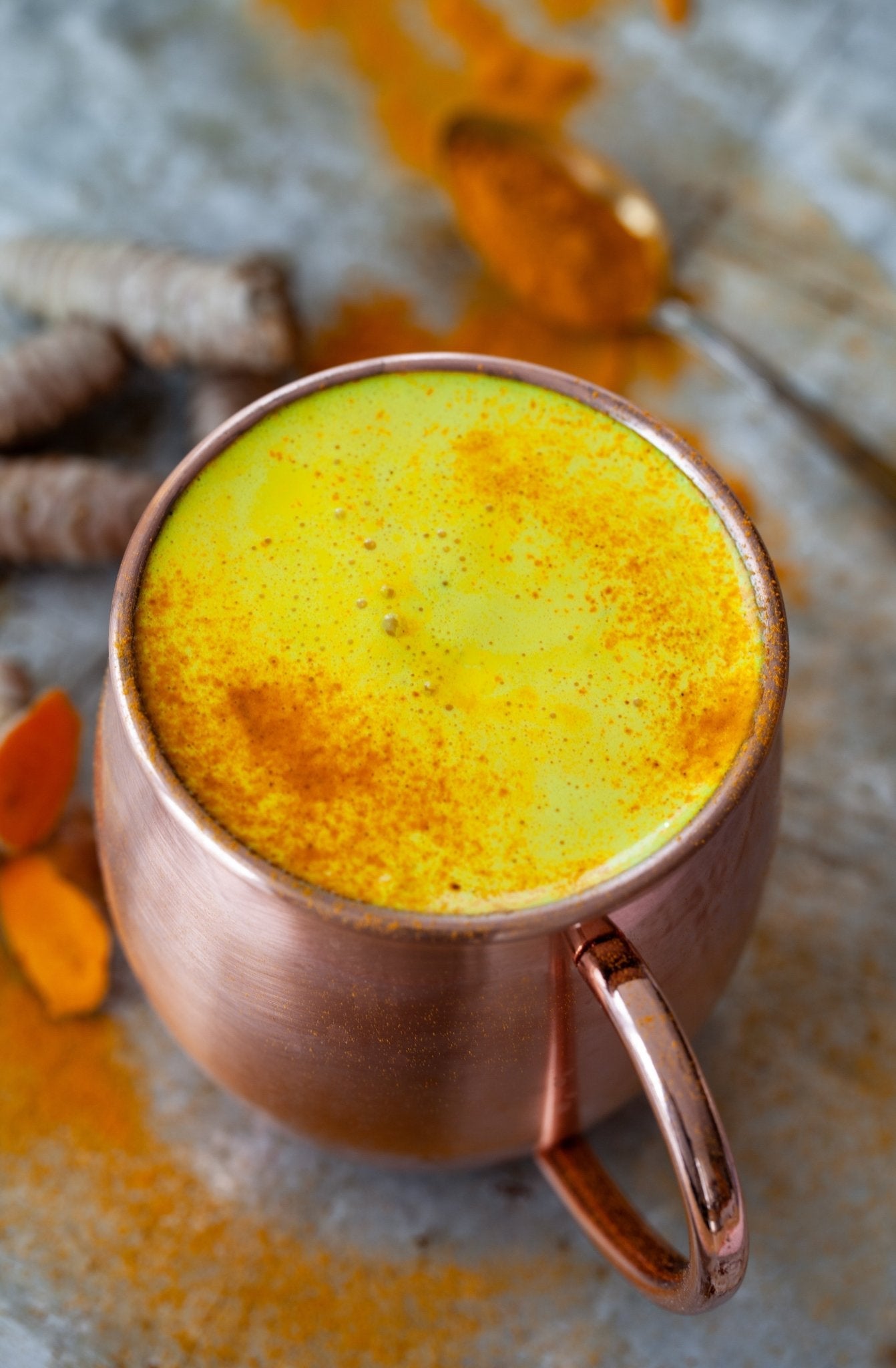 Copper mug filled with a yellow turmeric latte, surrounded by turmeric roots on a stone surface.