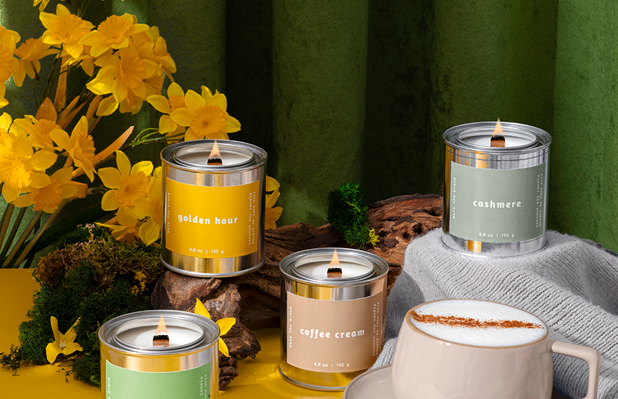 Candles with labels and a cup of coffee on a yellow surface with a green curtain background