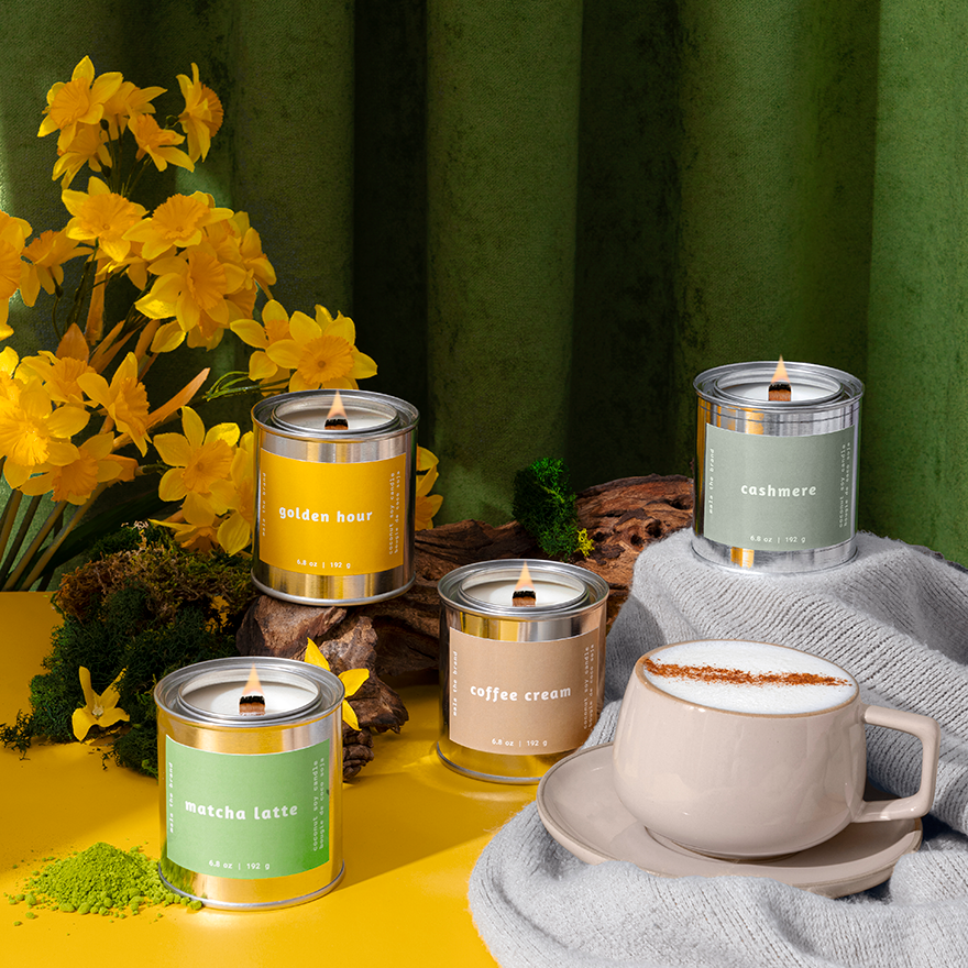 Candles with labels and a cup of coffee on a yellow surface with a green curtain background