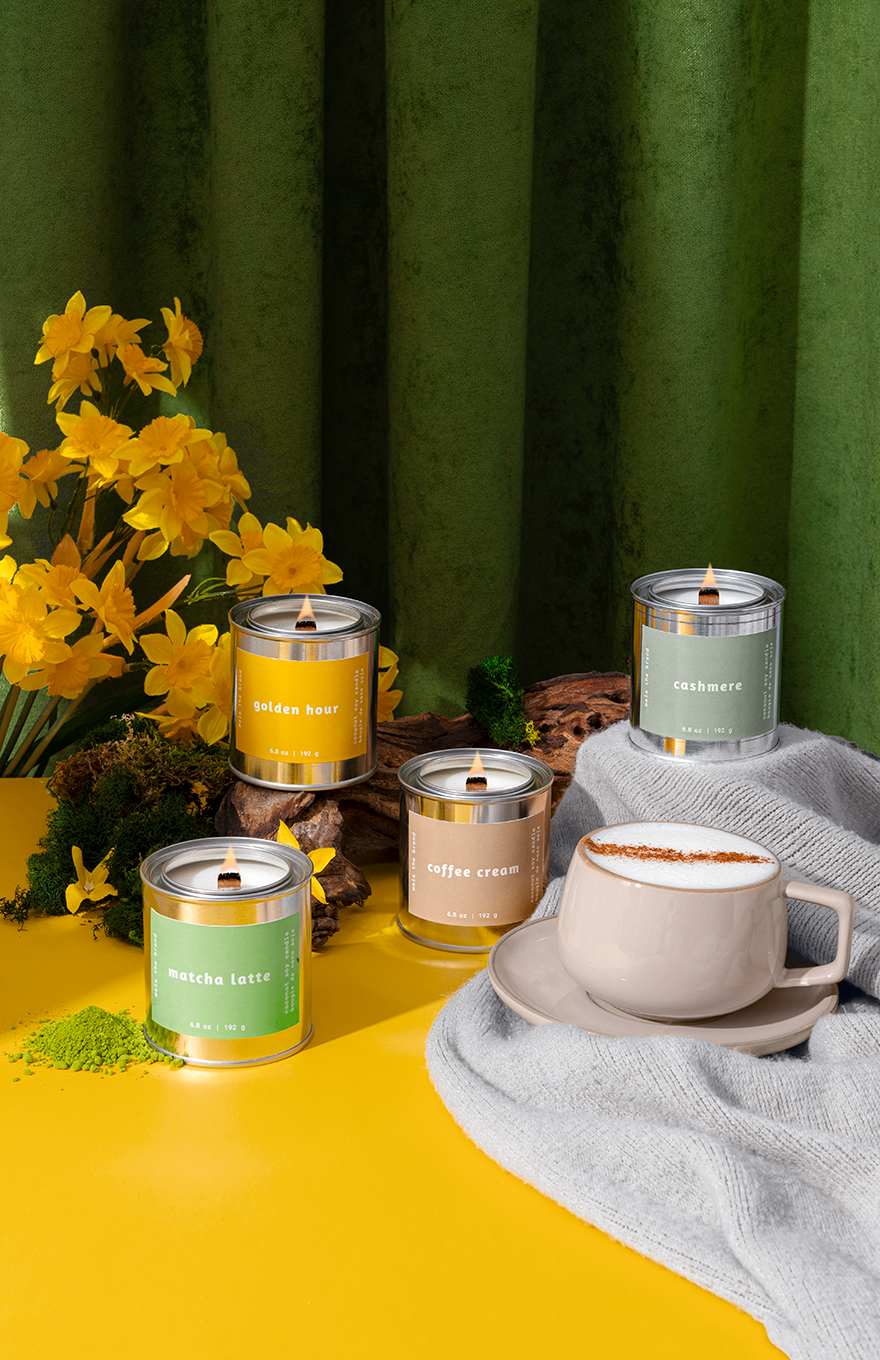 Candles with labels and a cup of coffee on a yellow surface with a green curtain background