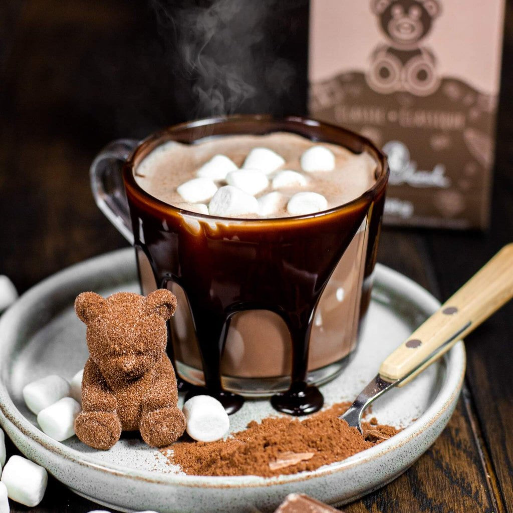 Hot chocolate with marshmallows, a small teddy bear, and a spoon on a wooden surface with a box in the background.