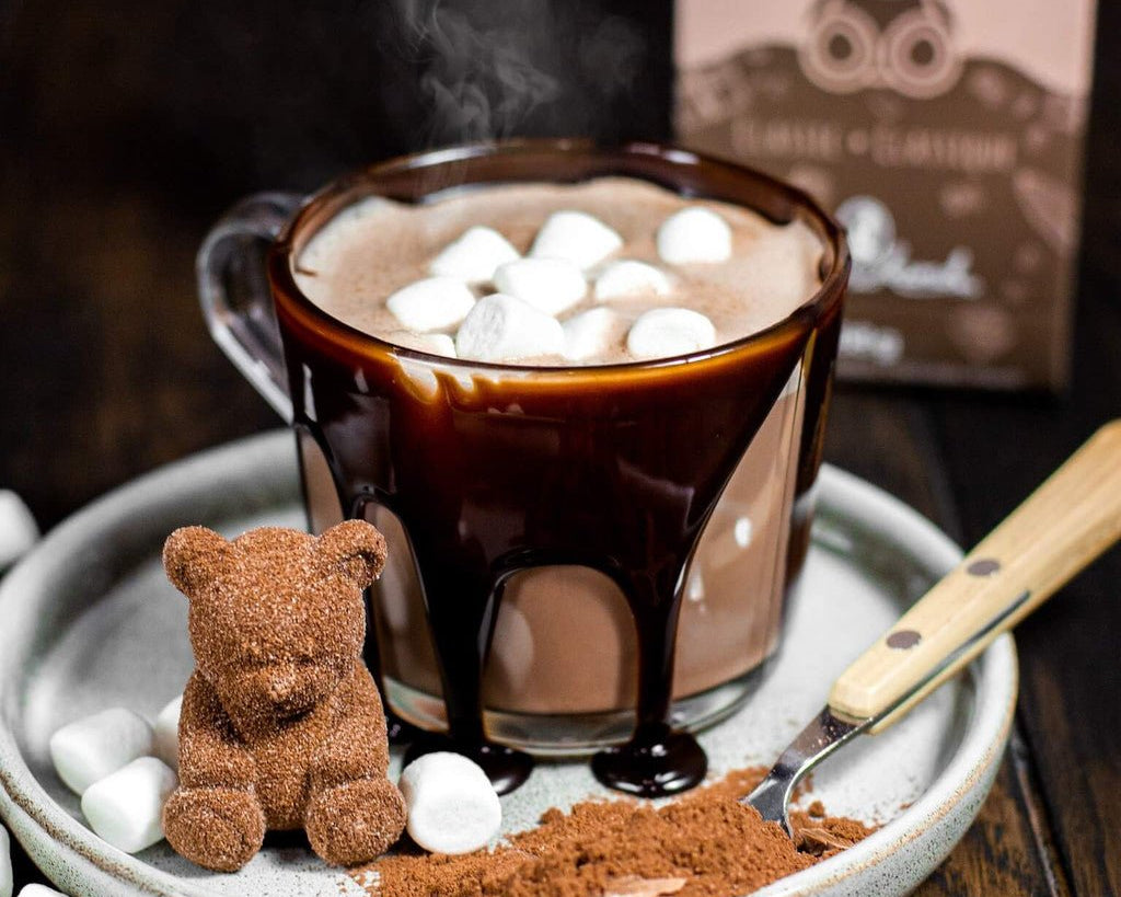 Hot chocolate with marshmallows, a small teddy bear, and a spoon on a wooden surface with a box in the background.