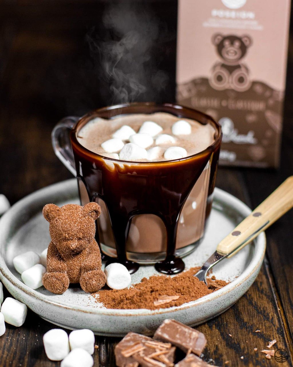 Hot chocolate with marshmallows, a small teddy bear, and a spoon on a wooden surface with a box in the background.