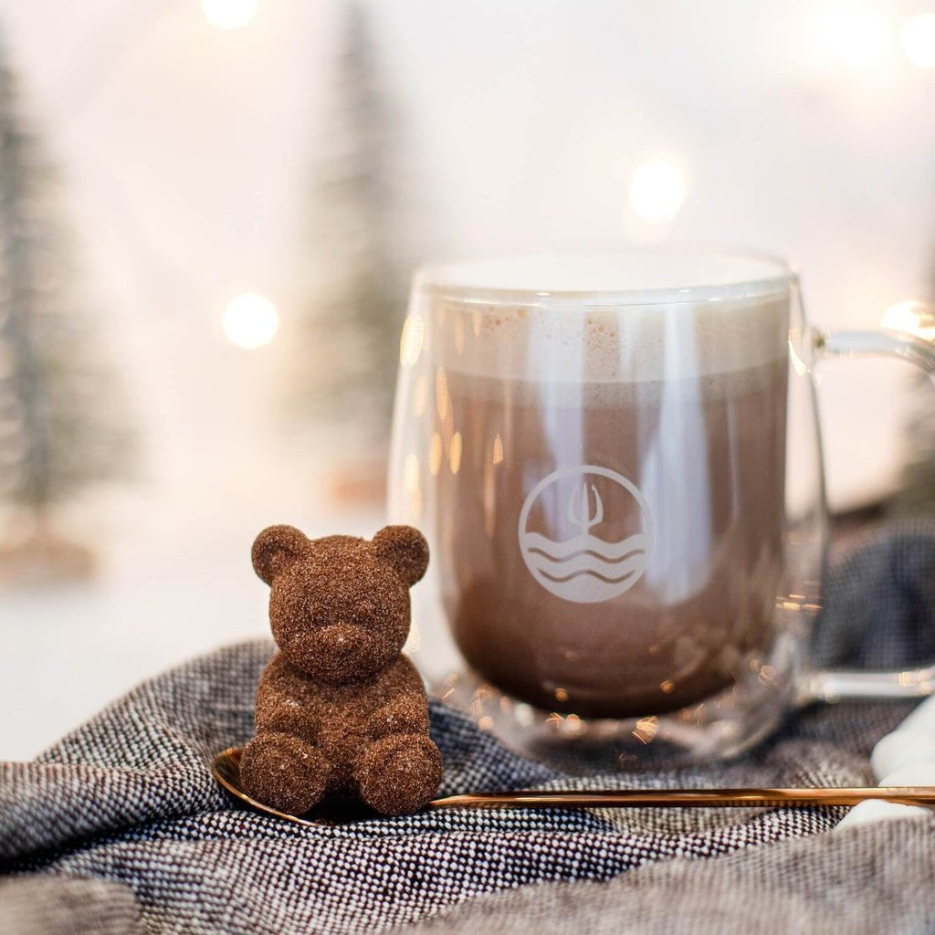 Small brown teddy bear next to a clear mug with a logo on a textured surface with a blurred natural background.