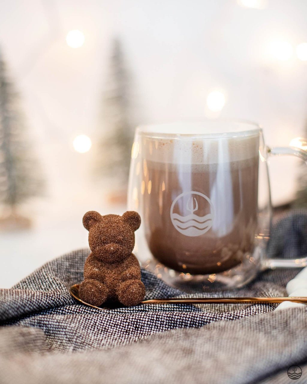 Small brown teddy bear next to a clear mug with a logo on a textured surface with a blurred natural background.