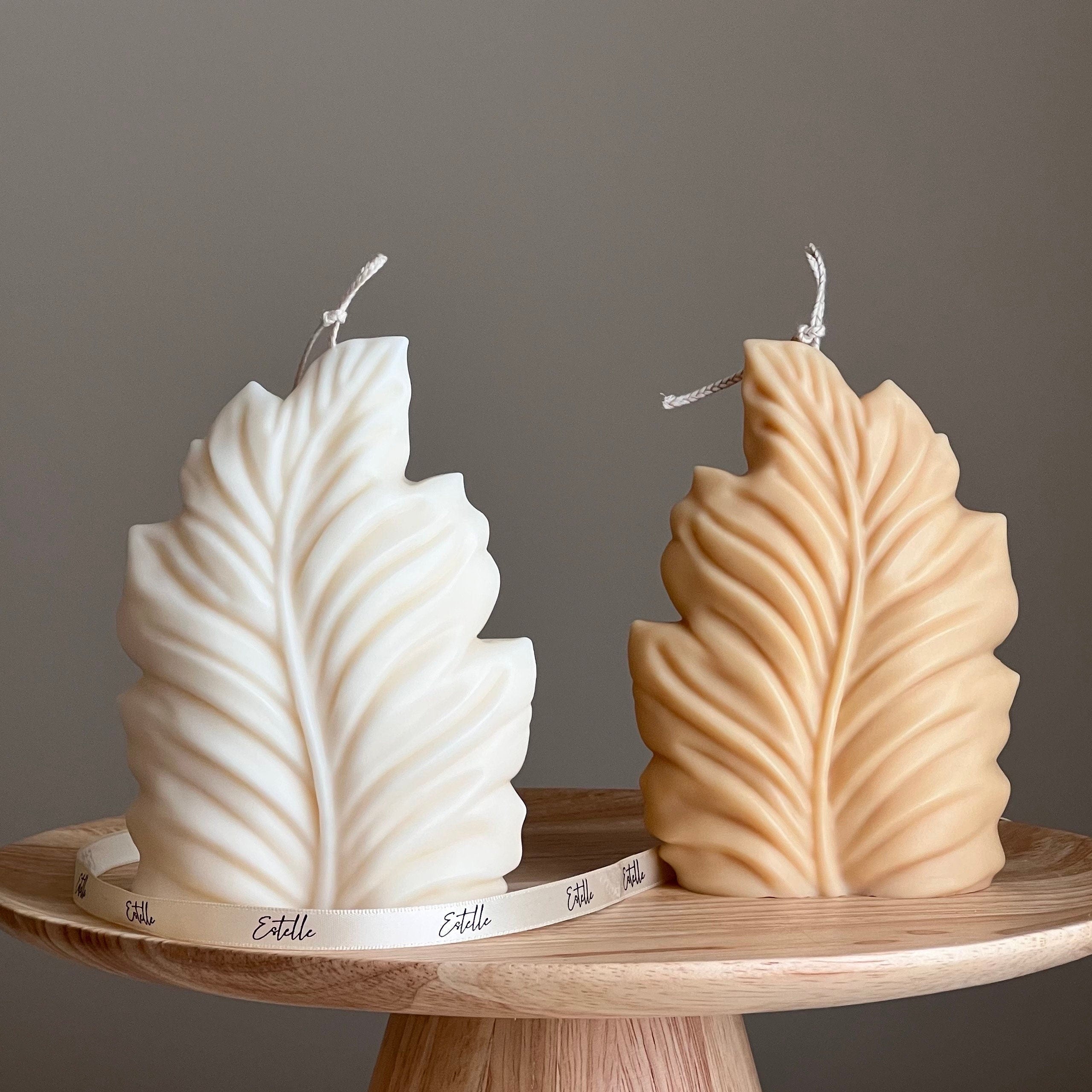 Two leaf-shaped candles, one white and one beige, on a wooden stand with a gray background.