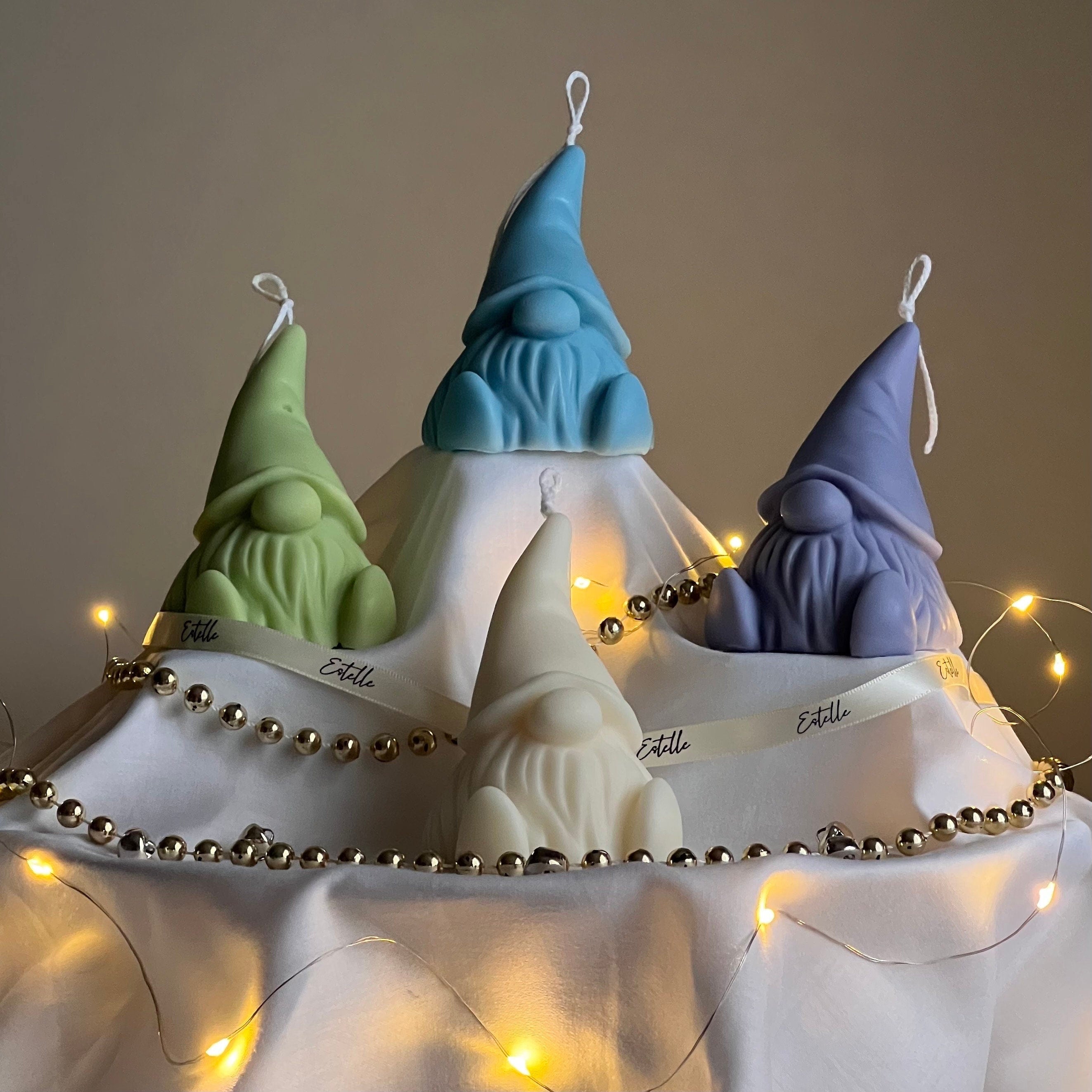 Four gnome-shaped candles with blue, green, white, and purple hats on a white cloth with fairy lights.