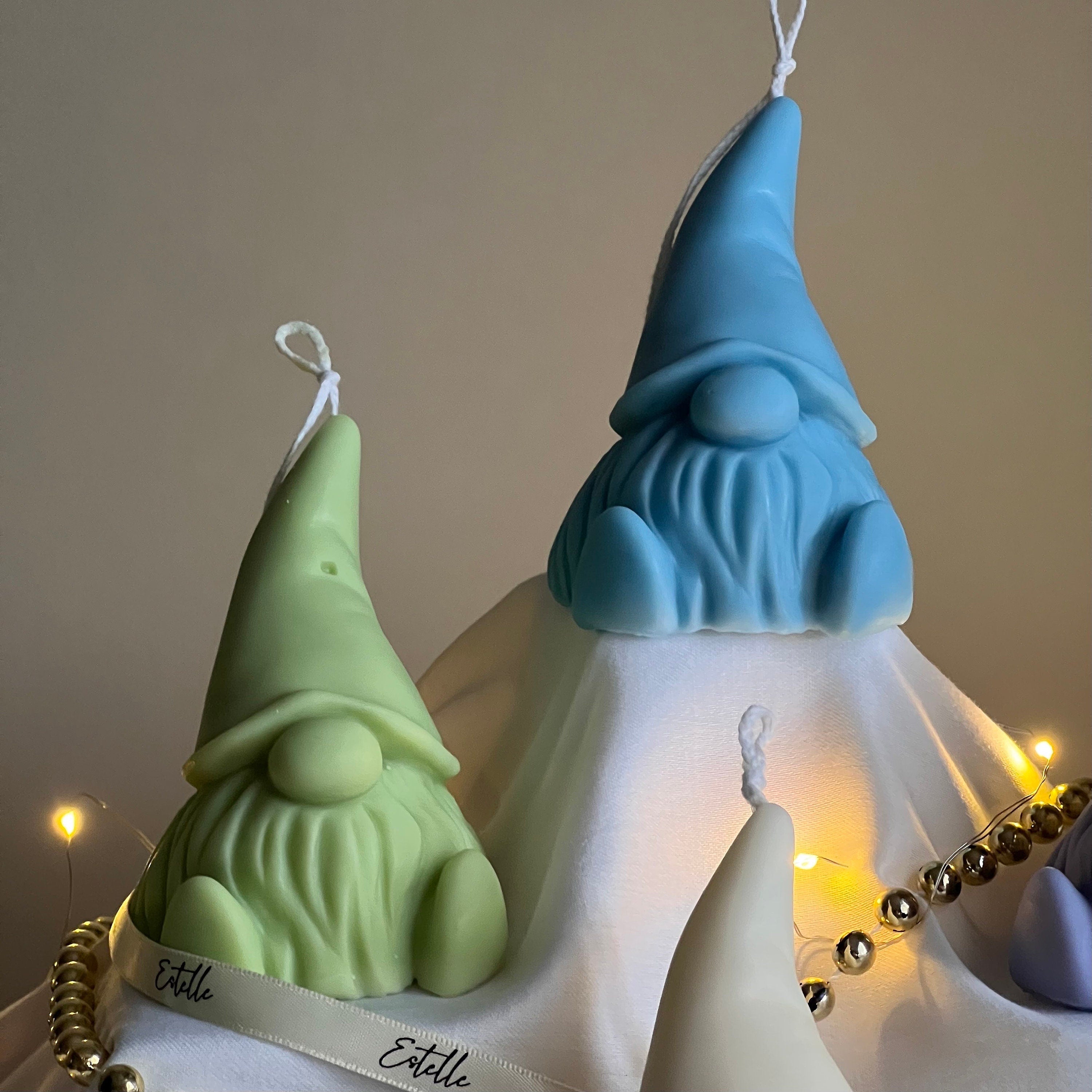 Three gnome-shaped candles in green, blue, and white with 'Estelle' branding on a neutral background.