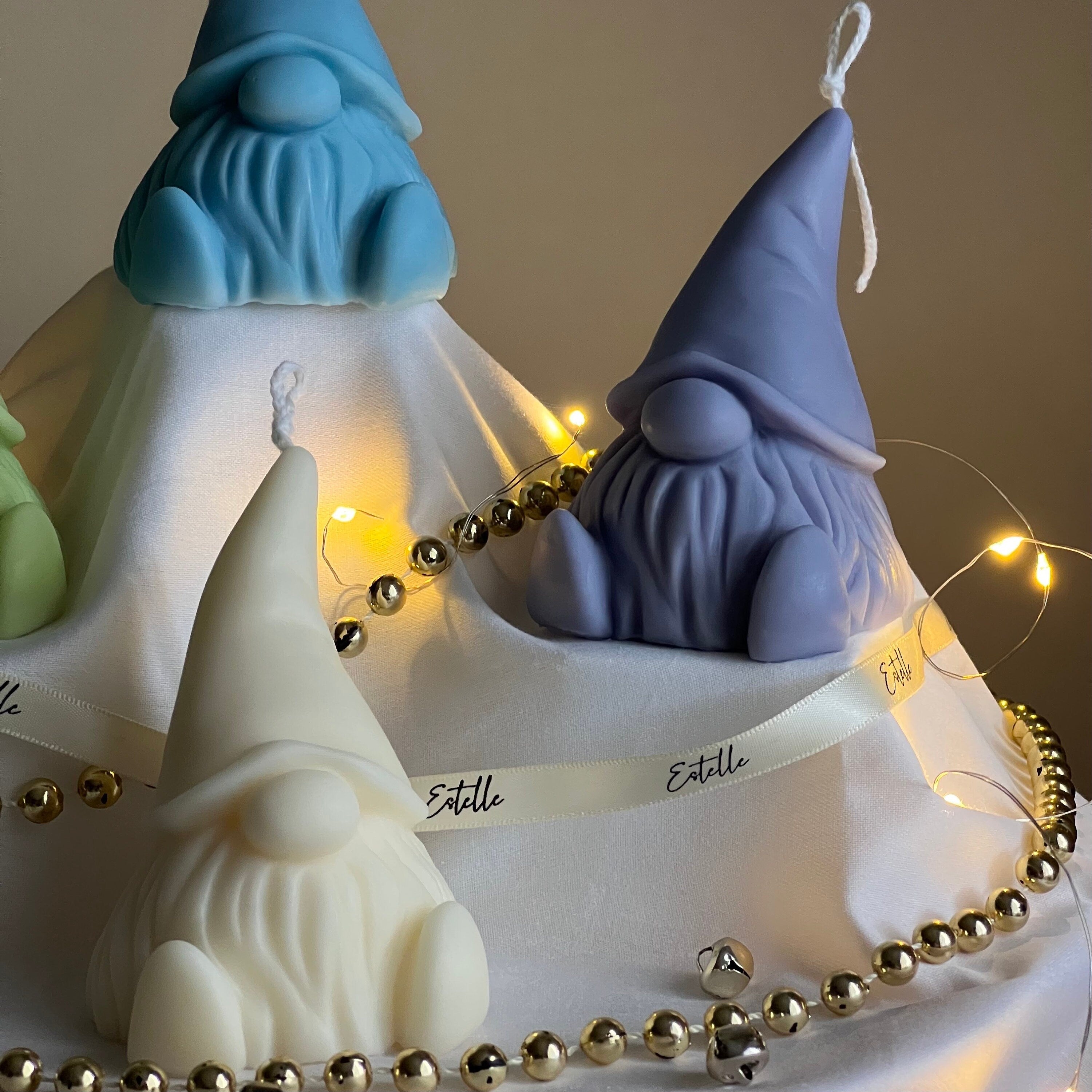 Three gnome-shaped candles with blue, purple, and white hats on a white base with gold beads and string lights.