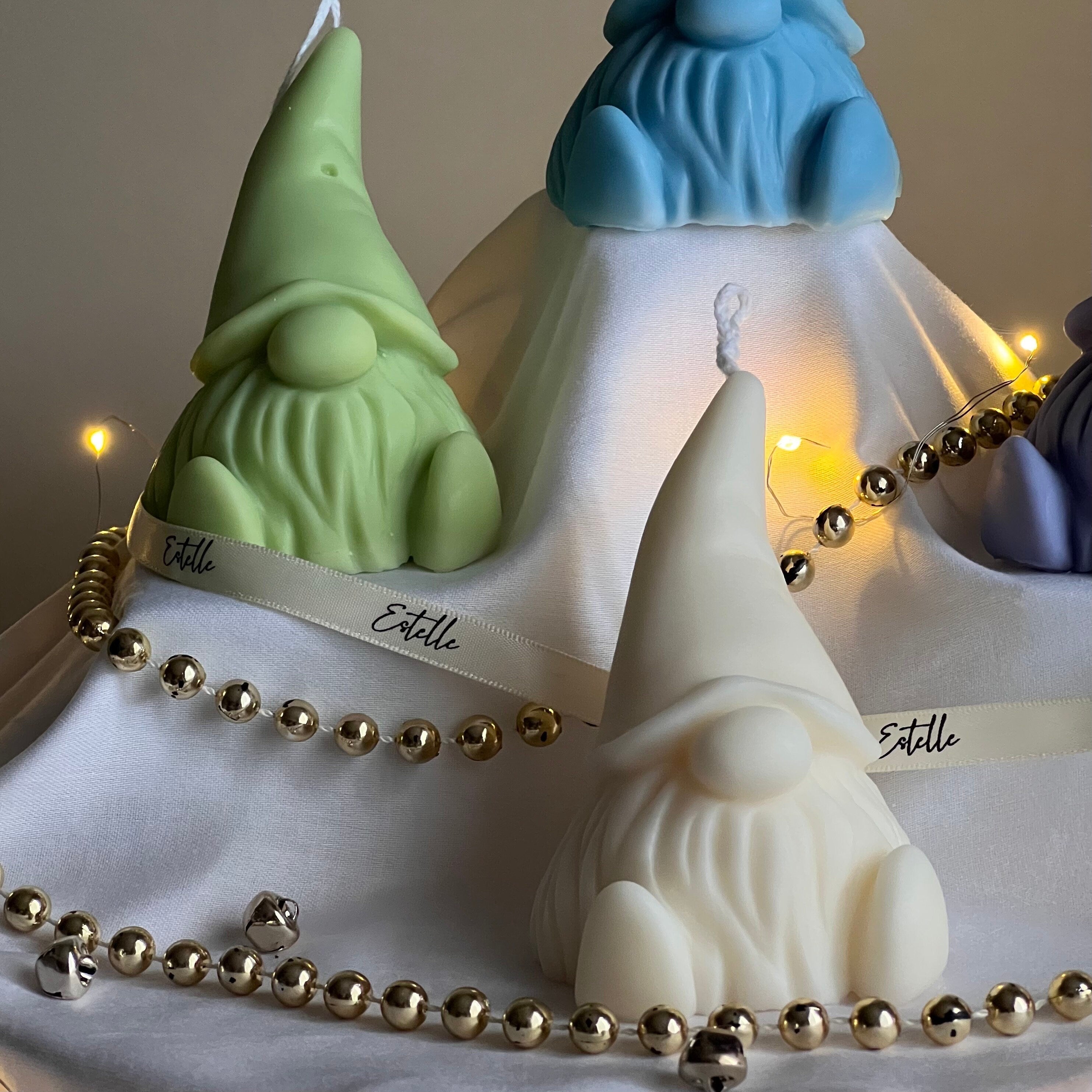 Three gnome-shaped candles with different colors and a string of lights on a white surface.
