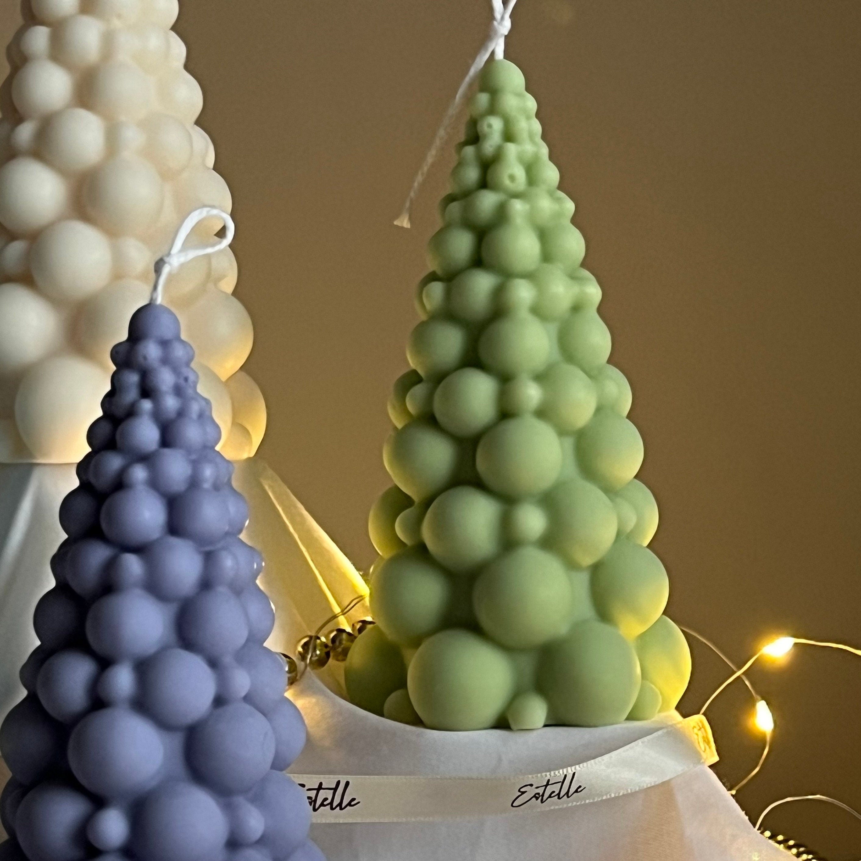 Three bubble-textured candles in purple, green, and white on a fabric with 'Estelle' branding, against a warm-toned background.