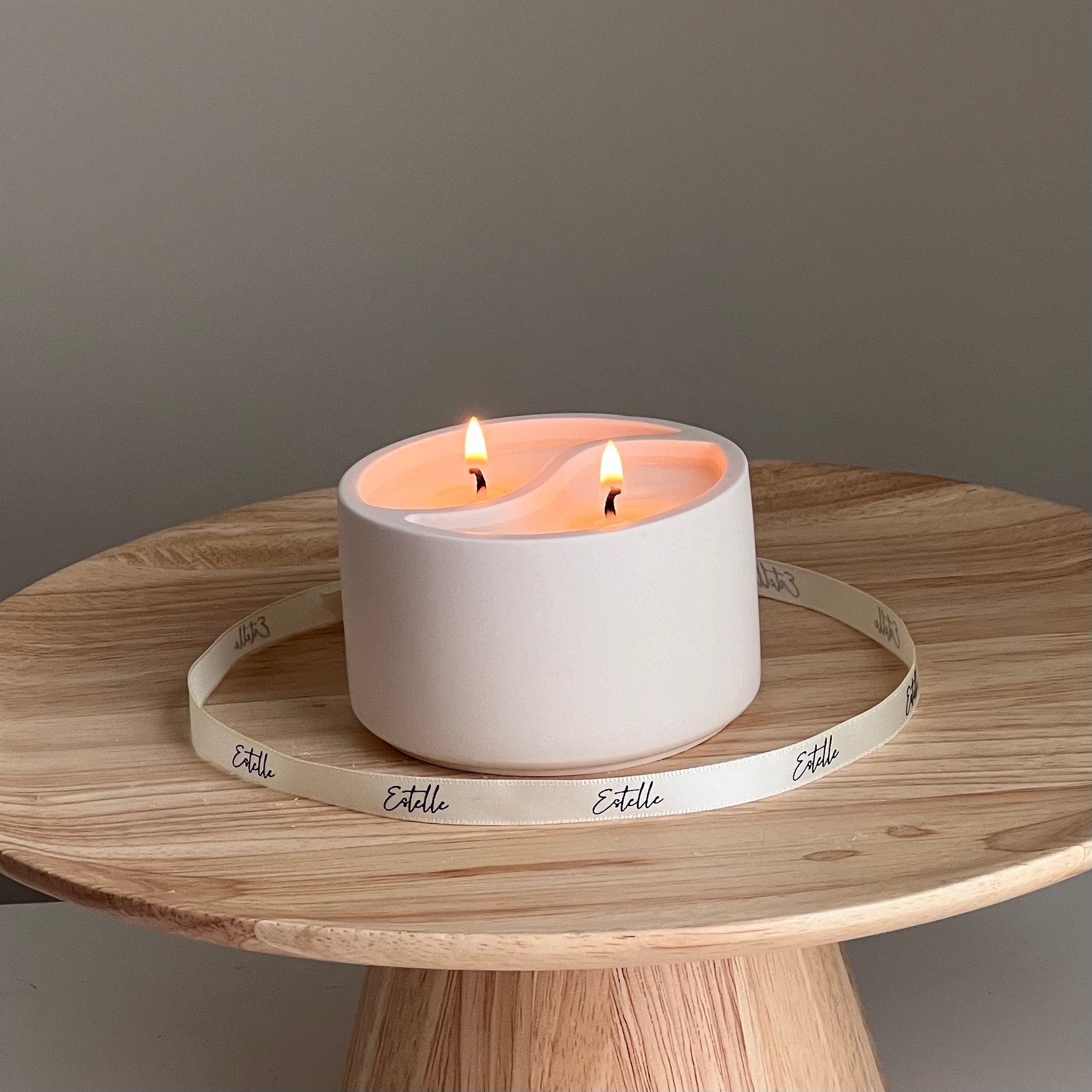 White candle holder with two lit candles on a wooden surface, featuring the brand 'Estelle'.