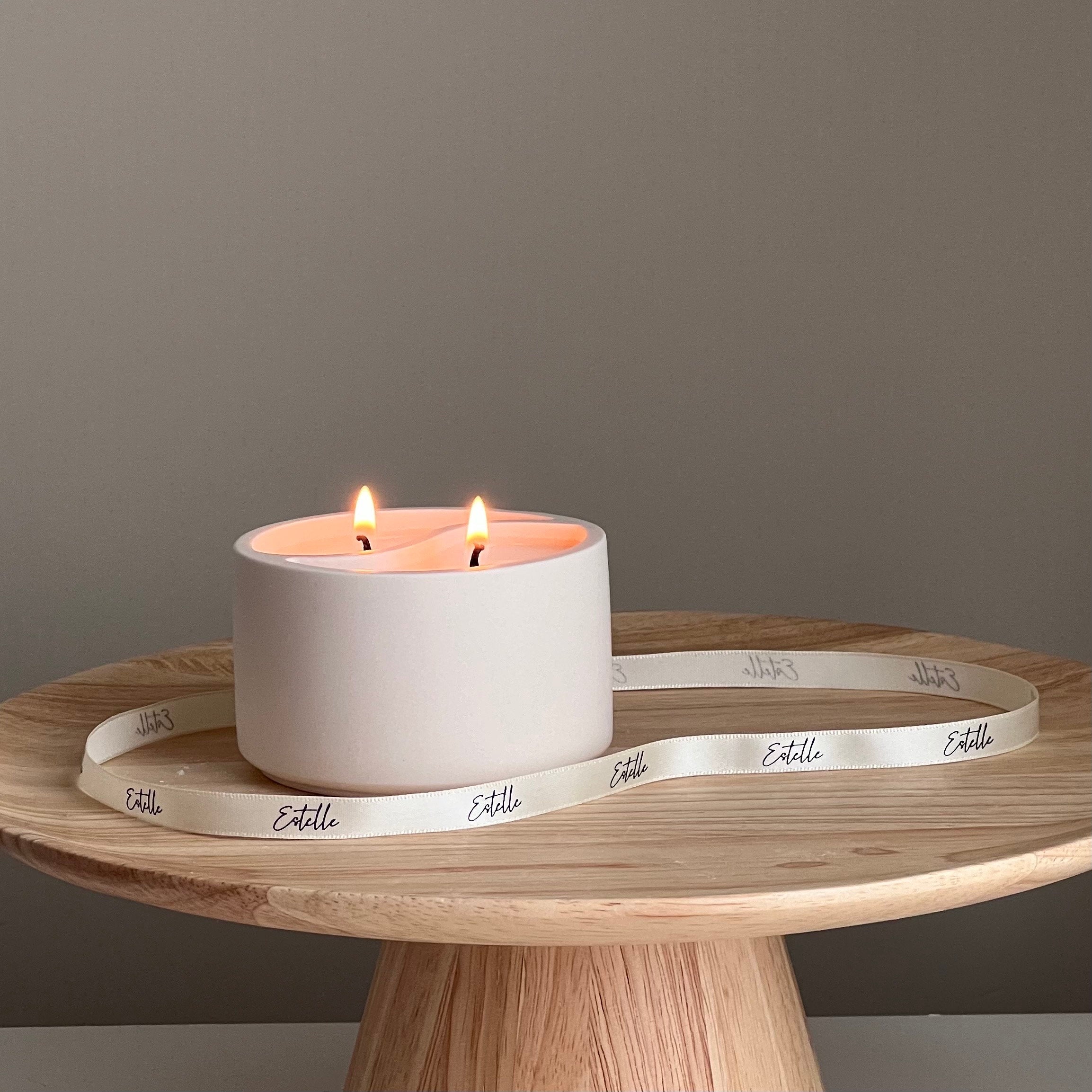 White candle in a pot with two flames on a wooden surface, surrounded by 'Estelle' branded ribbon.