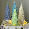 Four tree-shaped candles in blue, green, and white on a gray surface with string lights.