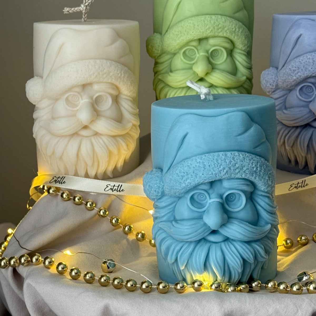 Four candles with Santa Claus faces on a gray surface with string lights from Estelle Studios