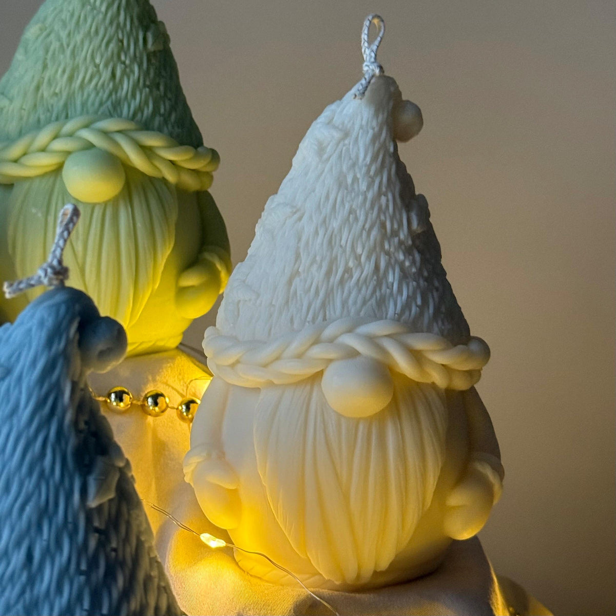 Three gnome-shaped candles with braided hair, one glowing, on a reflective surface.