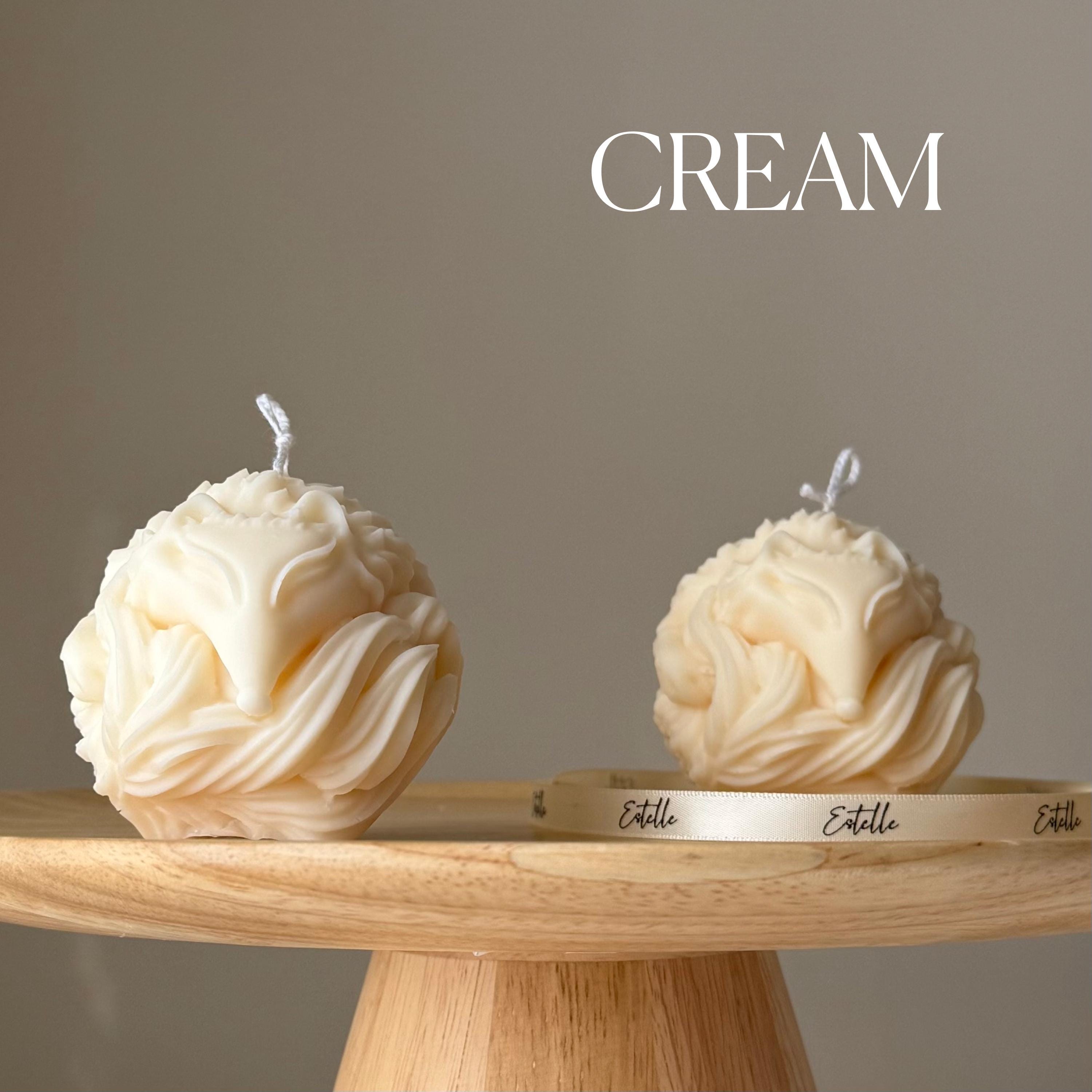 Two cream-colored spherical candles on a wooden stand with 'Estelle' branding against a neutral background.