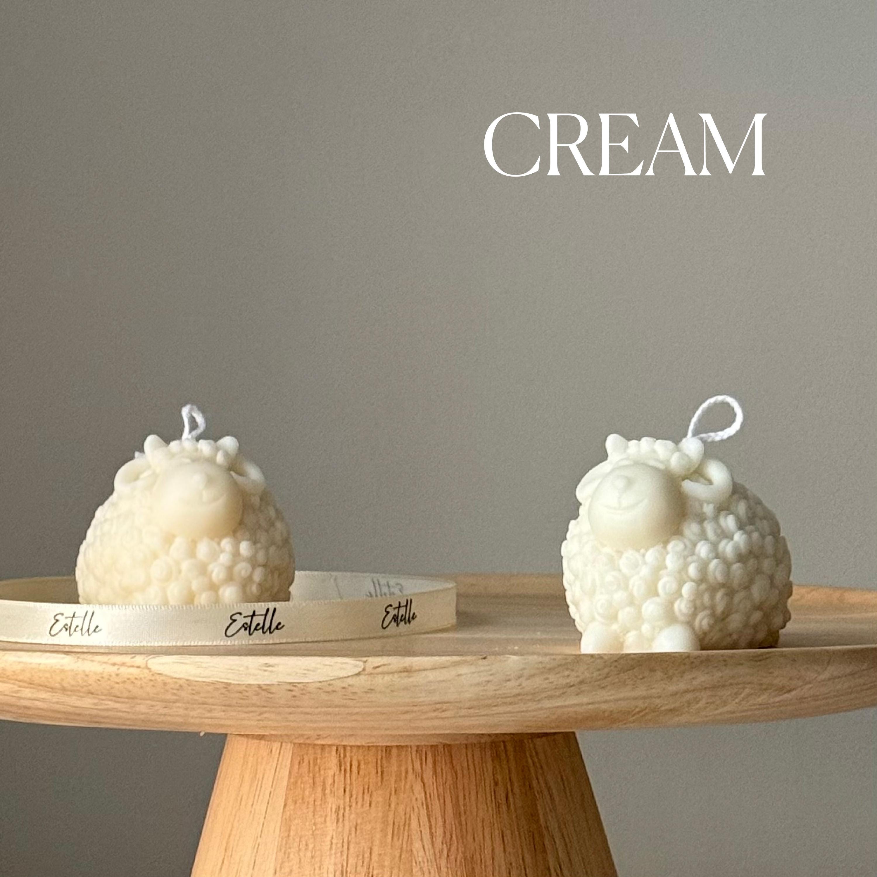 Two cream-colored bubble-textured sheep candles on a wooden stand with a gray background and 'CREAM' text.
