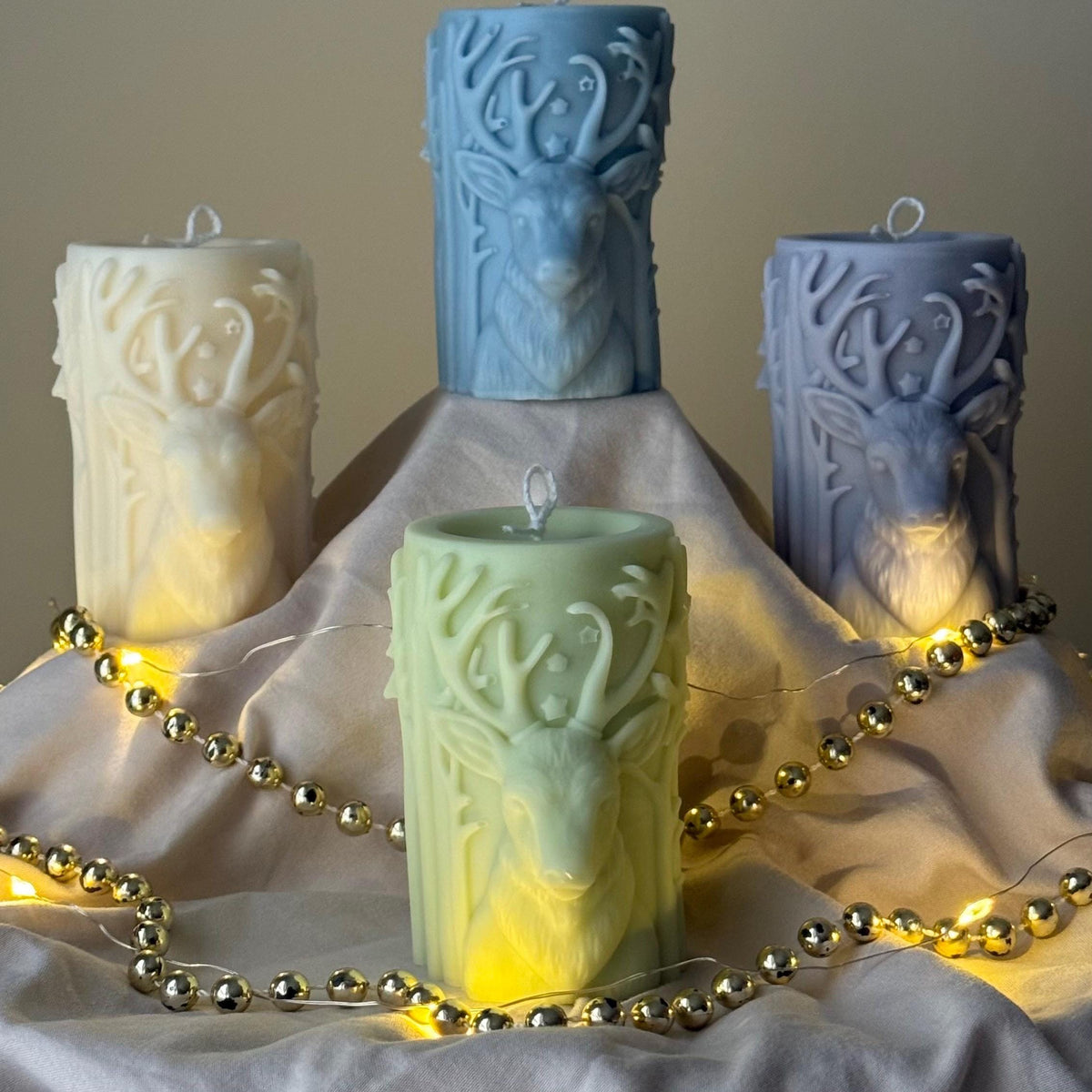 Decorative candles with deer head designs on a soft surface with string lights.