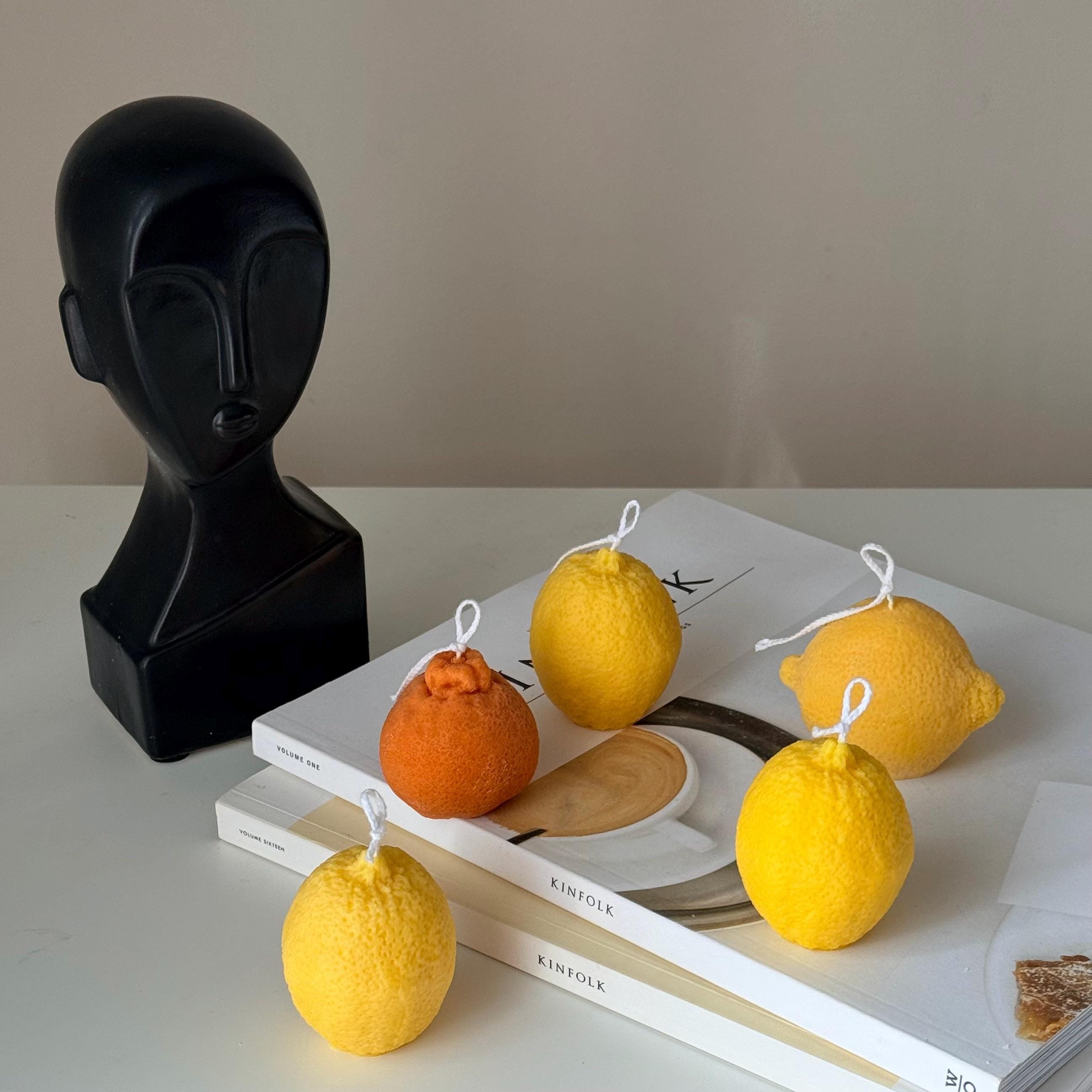 Decorative candles shaped like lemons and an orange on books with a black abstract sculpture in the background.
