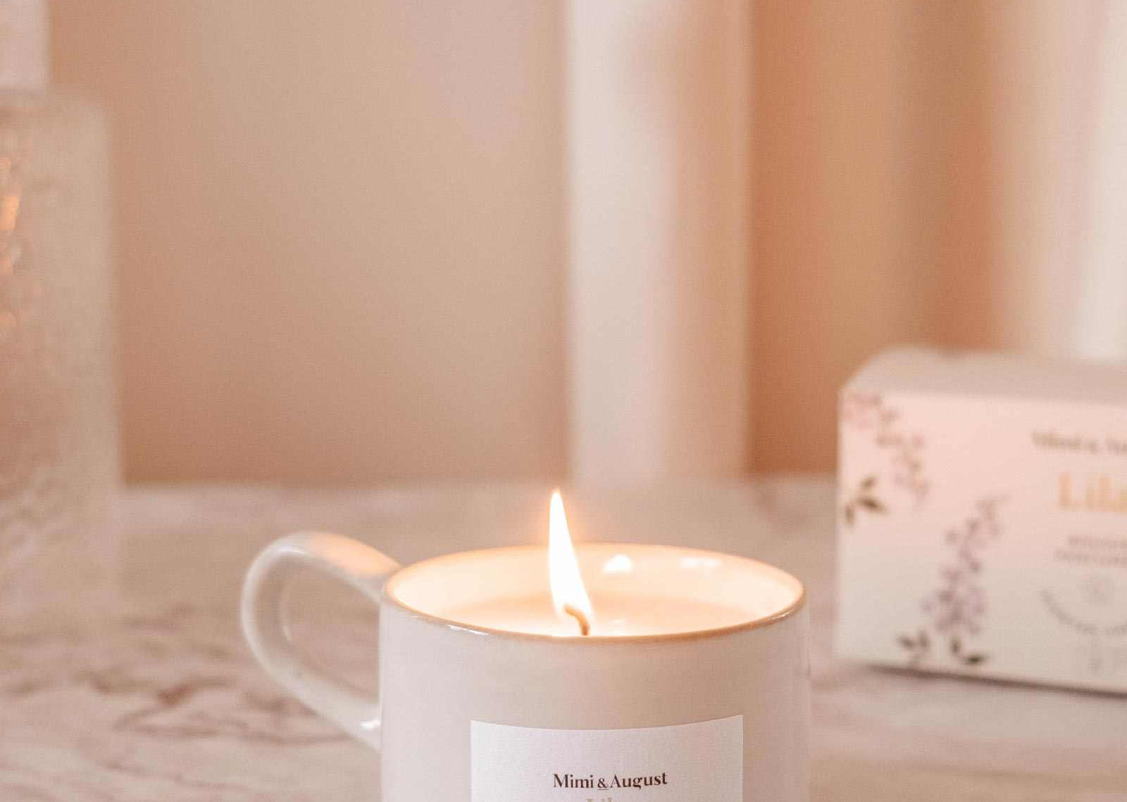 Lit candle in a mug with a visible label on a soft pink background mimi-and-august