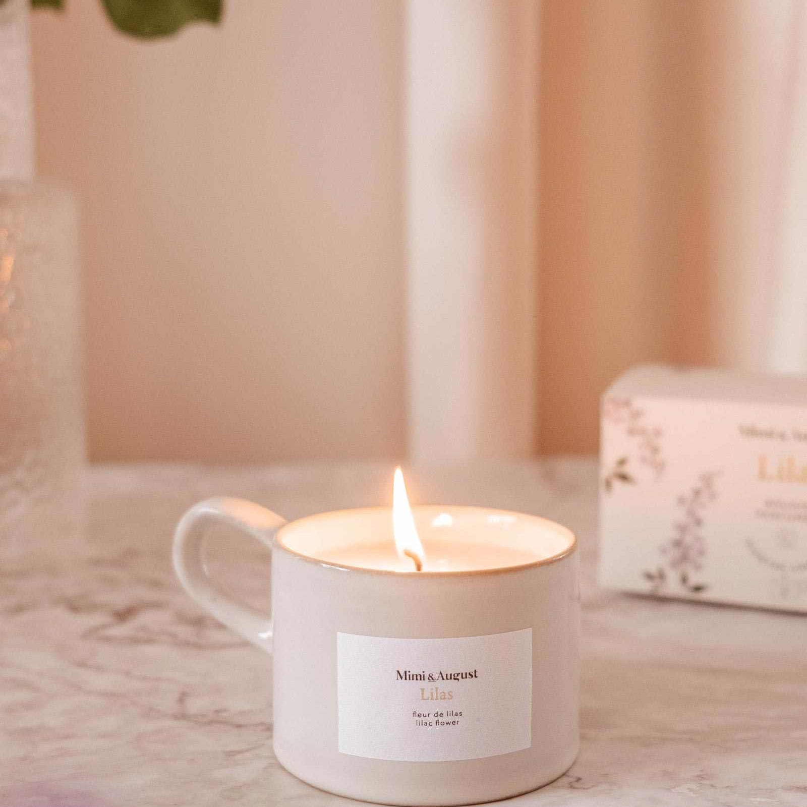 Lit candle in a mug with a visible label on a soft pink background mimi-and-august