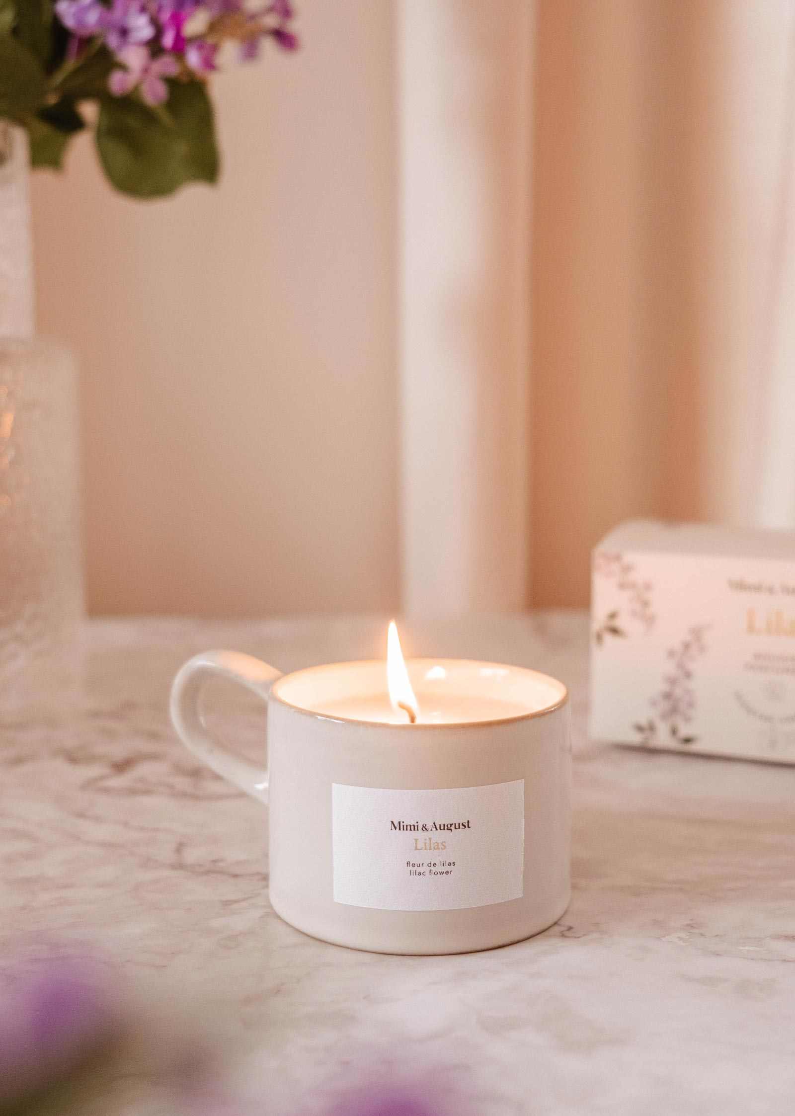 Lit candle in a mug with a visible label on a soft pink background mimi-and-august